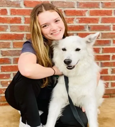 Olivia holding onto a white dog and smiling in front of a brick wall. Olivia holding onto a white dog and smiling in front of a brick wall.