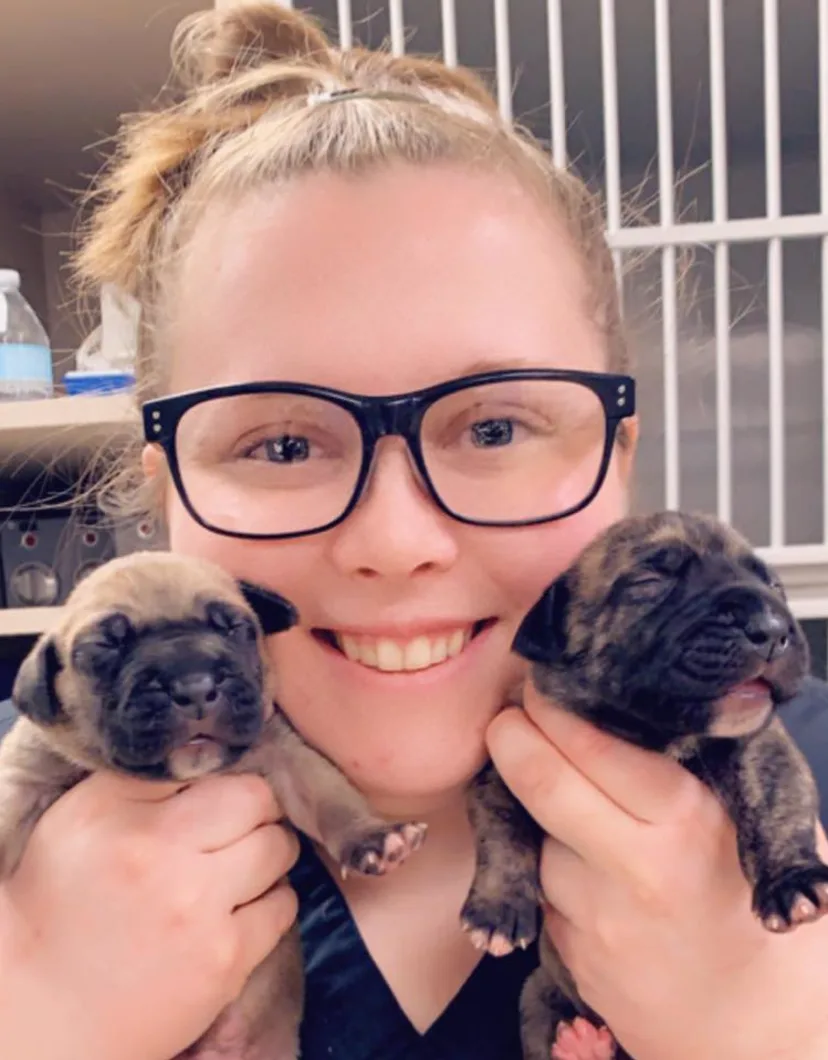 Kelsey's staff photo from University West Pet Clinic where she is holding two puppies in front of her face.