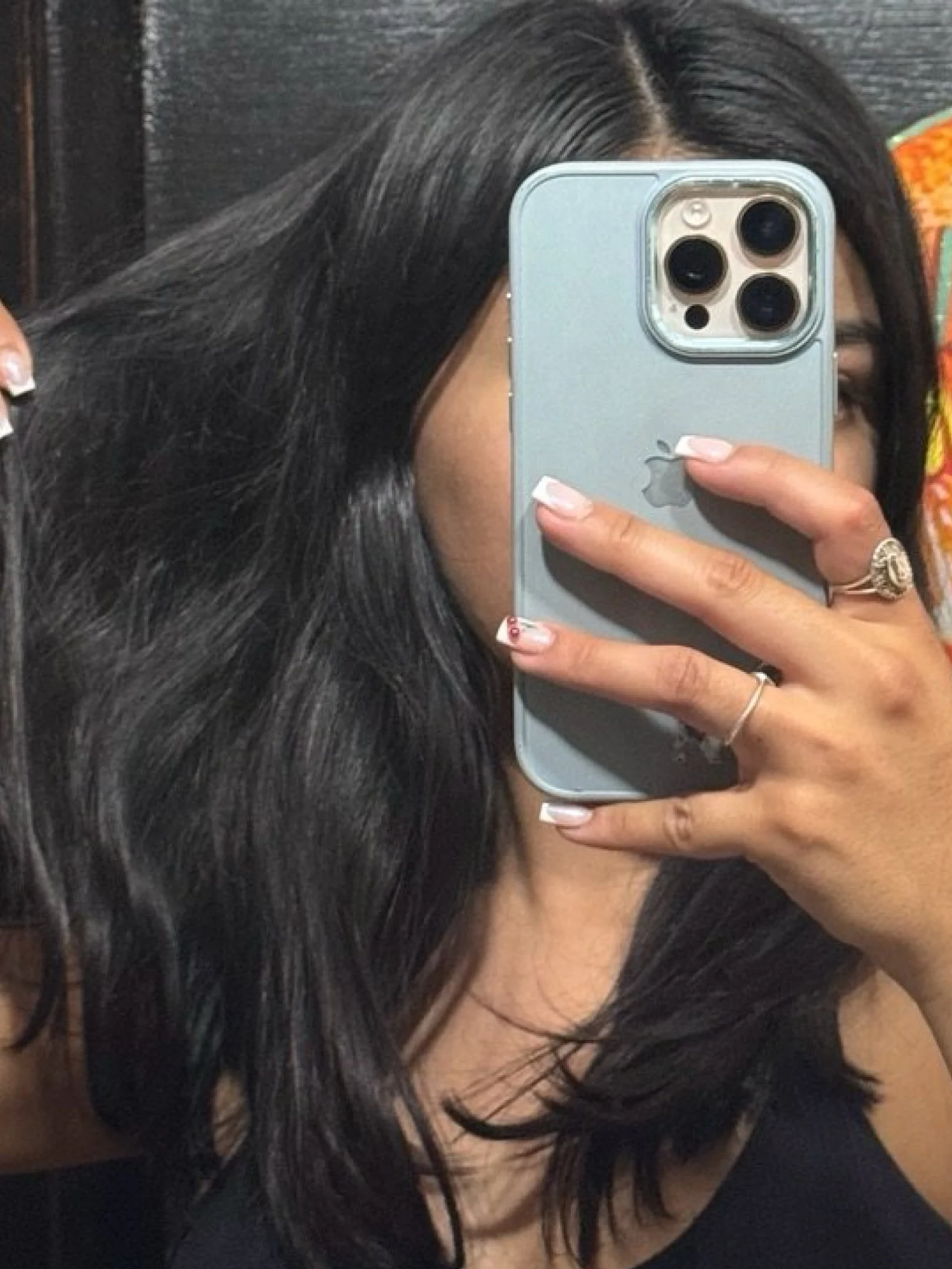 Woman with shoulder-length, sleek black hair taking a mirror selfie, partially covering her face with a light blue phone.