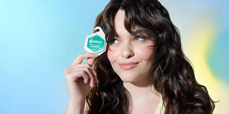 Smiling woman with dark wavy hair, holding a GEMZ shampoo package in her fingers, conveying the refreshing effect of GEMZ hair care.