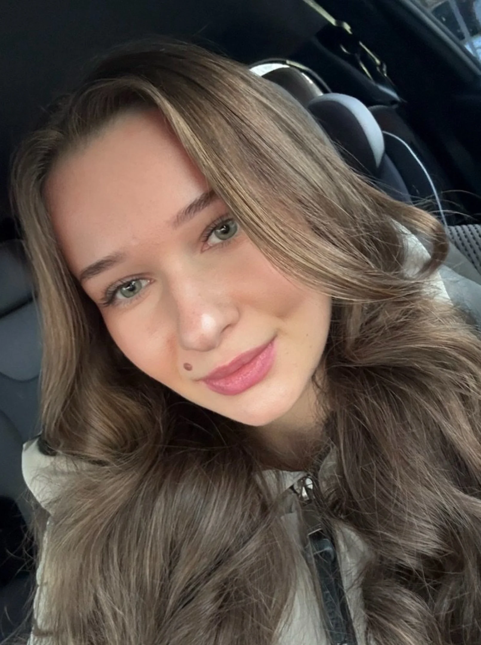 Young woman with long, wavy light brown hair, sitting in a car and smiling softly at the camera.