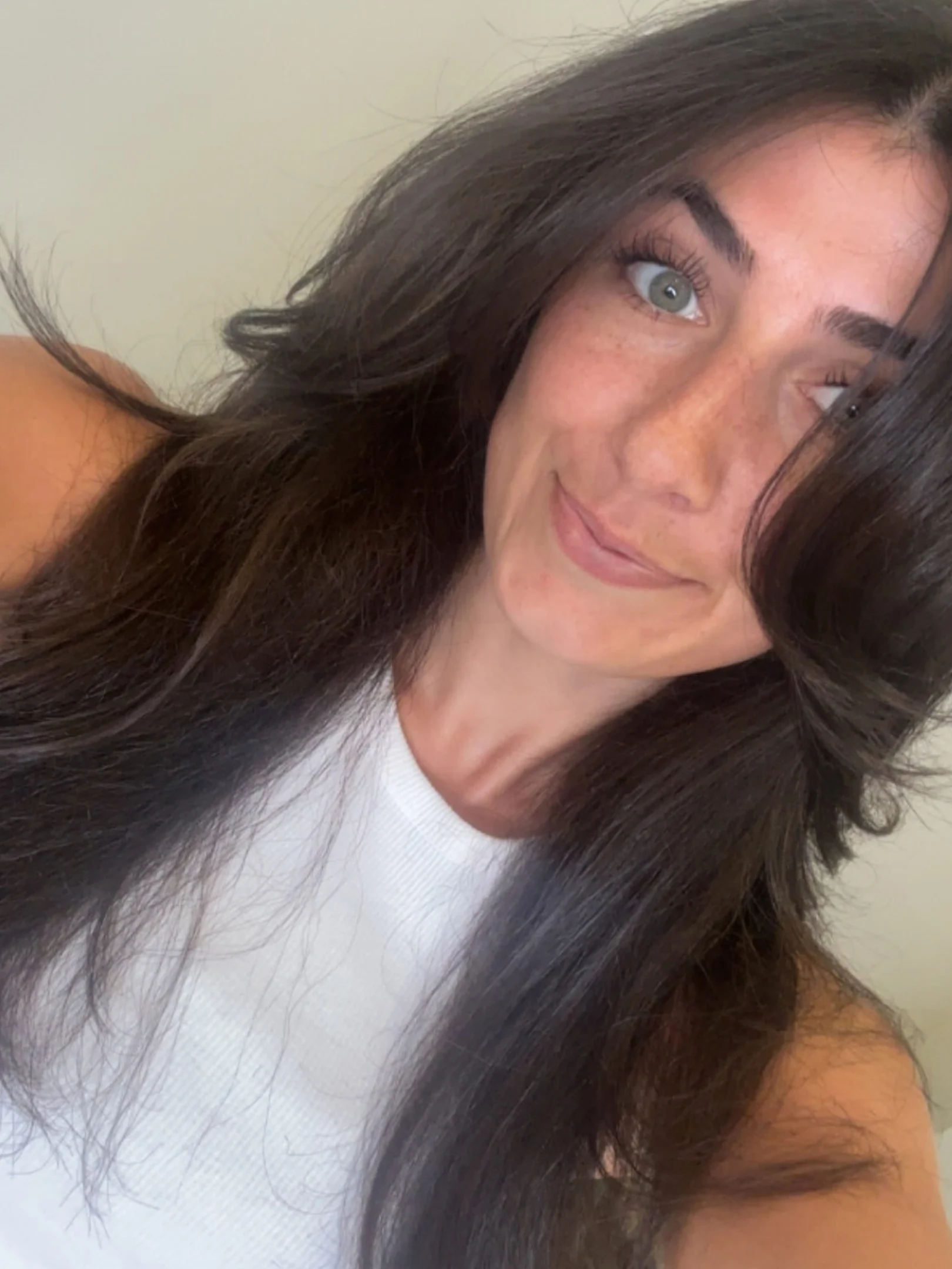 A woman with long dark brown hair and green eyes, wearing a white sleeveless top, smiling slightly at the camera in a well-lit setting.