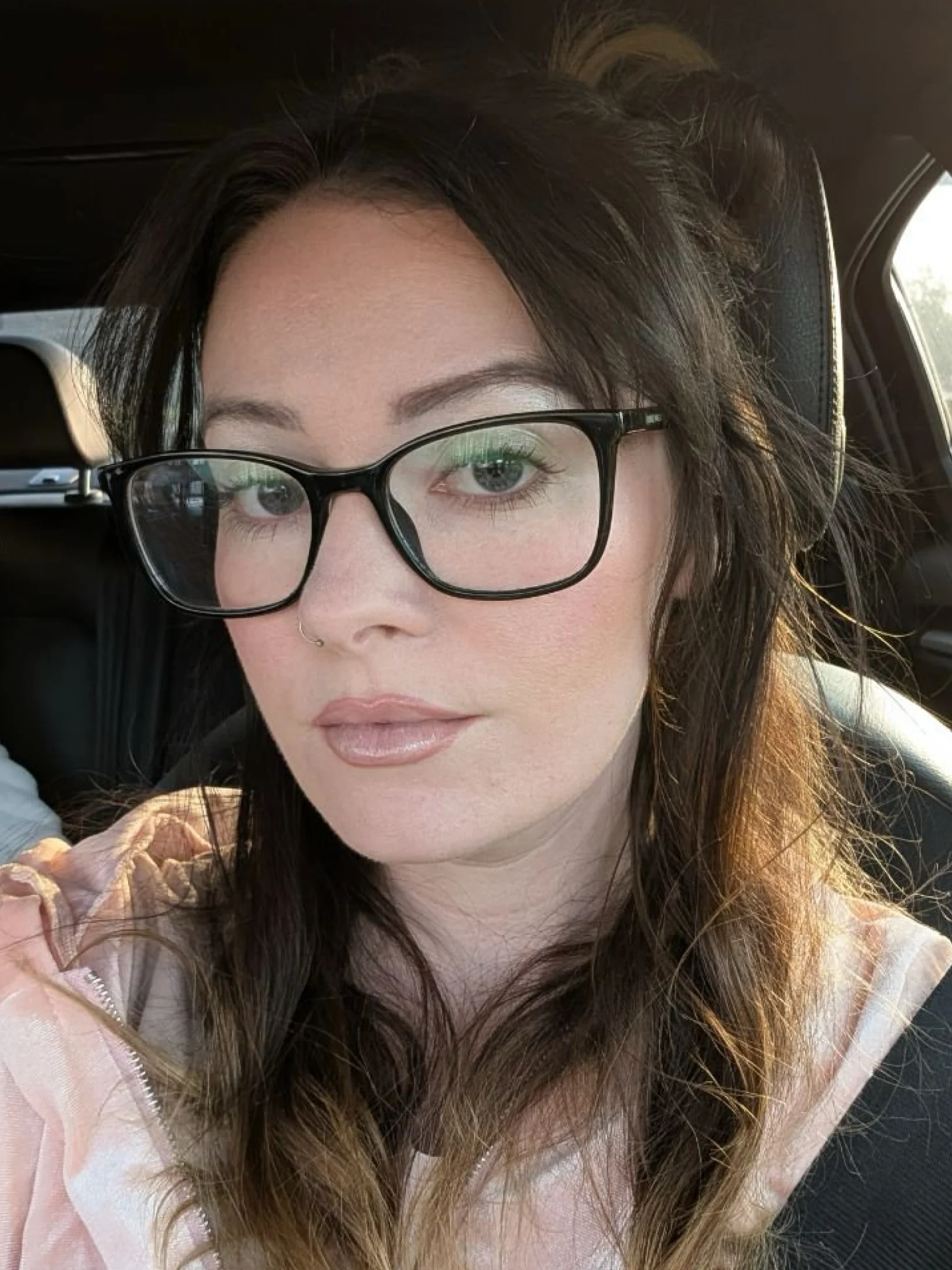 A woman with long dark hair, wearing black glasses and light makeup with green eyeshadow, sitting in a car and looking at the camera with a neutral expression.