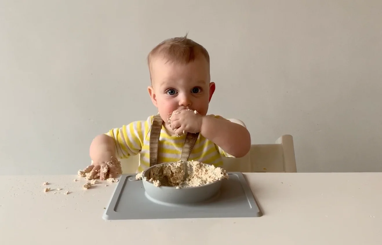 a photograph of a baby in a yellow onesie bringing a handful of ricotta to her mouth