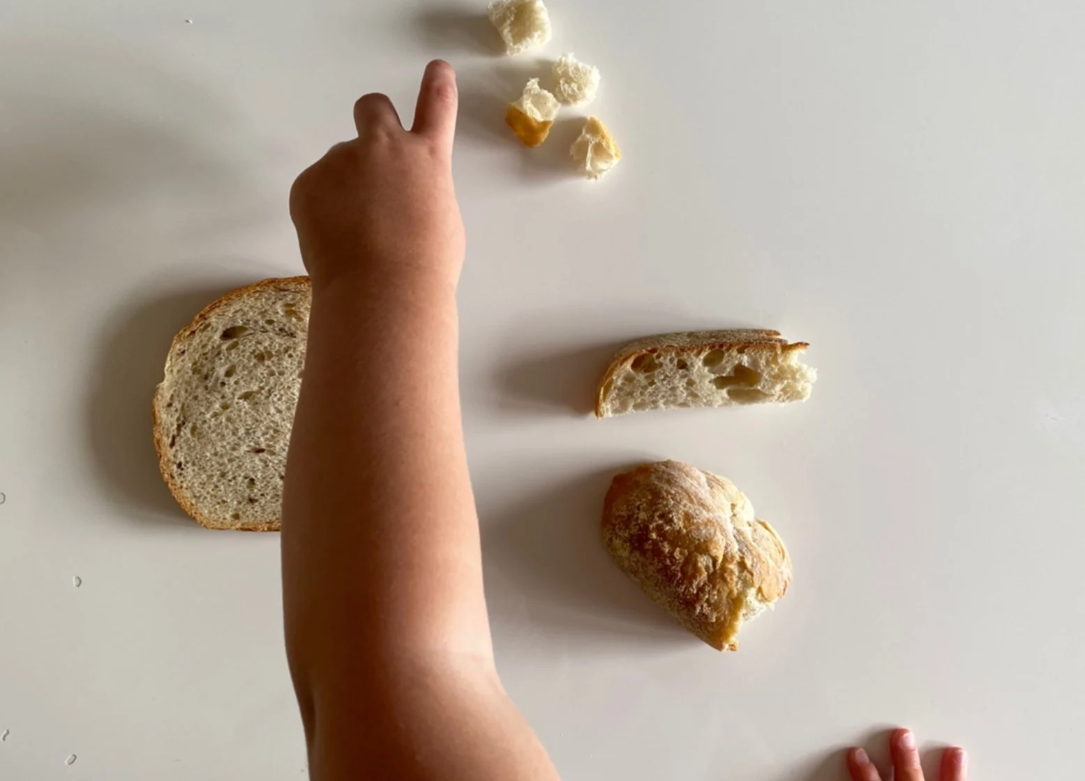 a toddler's arm reaching across a white table with three types of bread on it and pointing to bite-sized pieces of bread