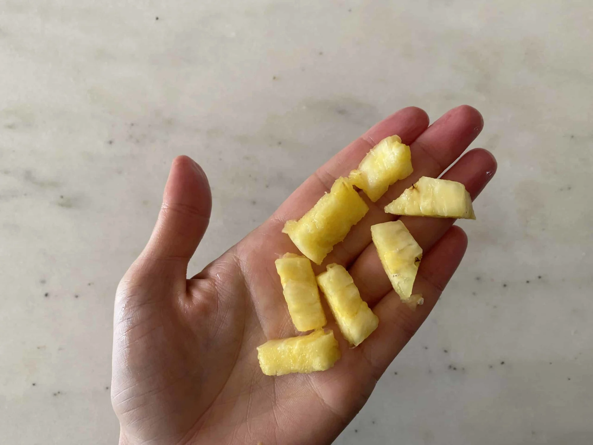 a hand holding seven bite-sized pieces of pineapple for babies 9 months+