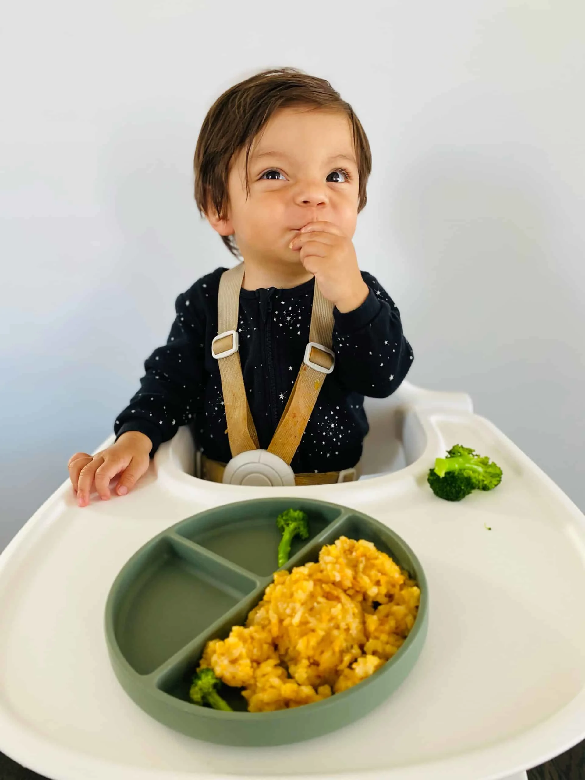 a baby in a highchair bringing a piece of food to his mouth with a plate of food in front of him