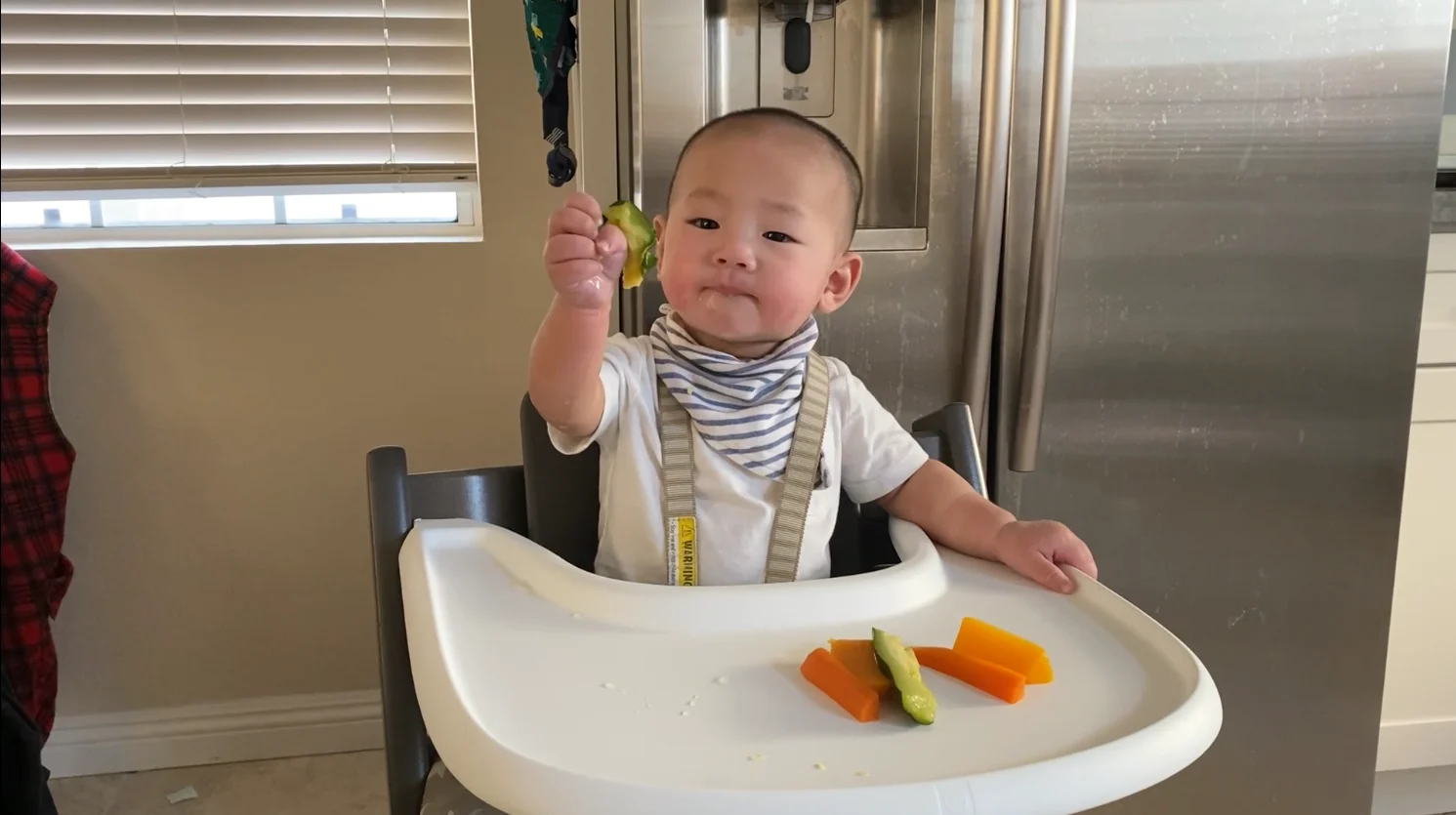 a photograph of a baby in a high chair with a few pieces of cooked vegetables on the tray in front of him and a piece of zucchini in his outstretched hand