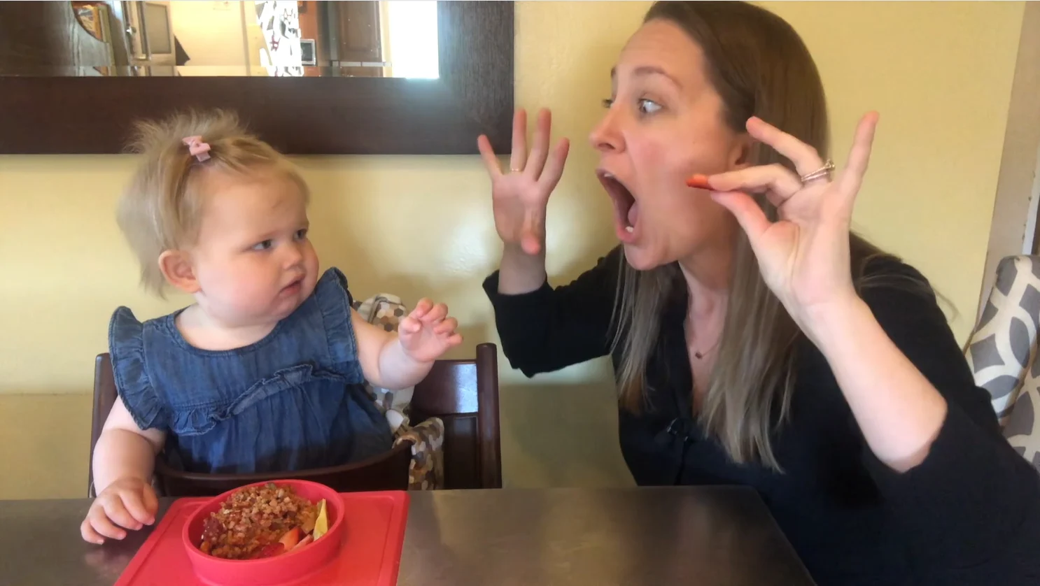 a feeding therapist sits next to a baby in a high chair at a table, with hands up and open to model her mouth chewing