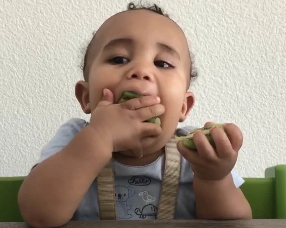 Un bebe se mete la mitad de un kiwi en la boca y sostiene la otra mitad en su otra mano