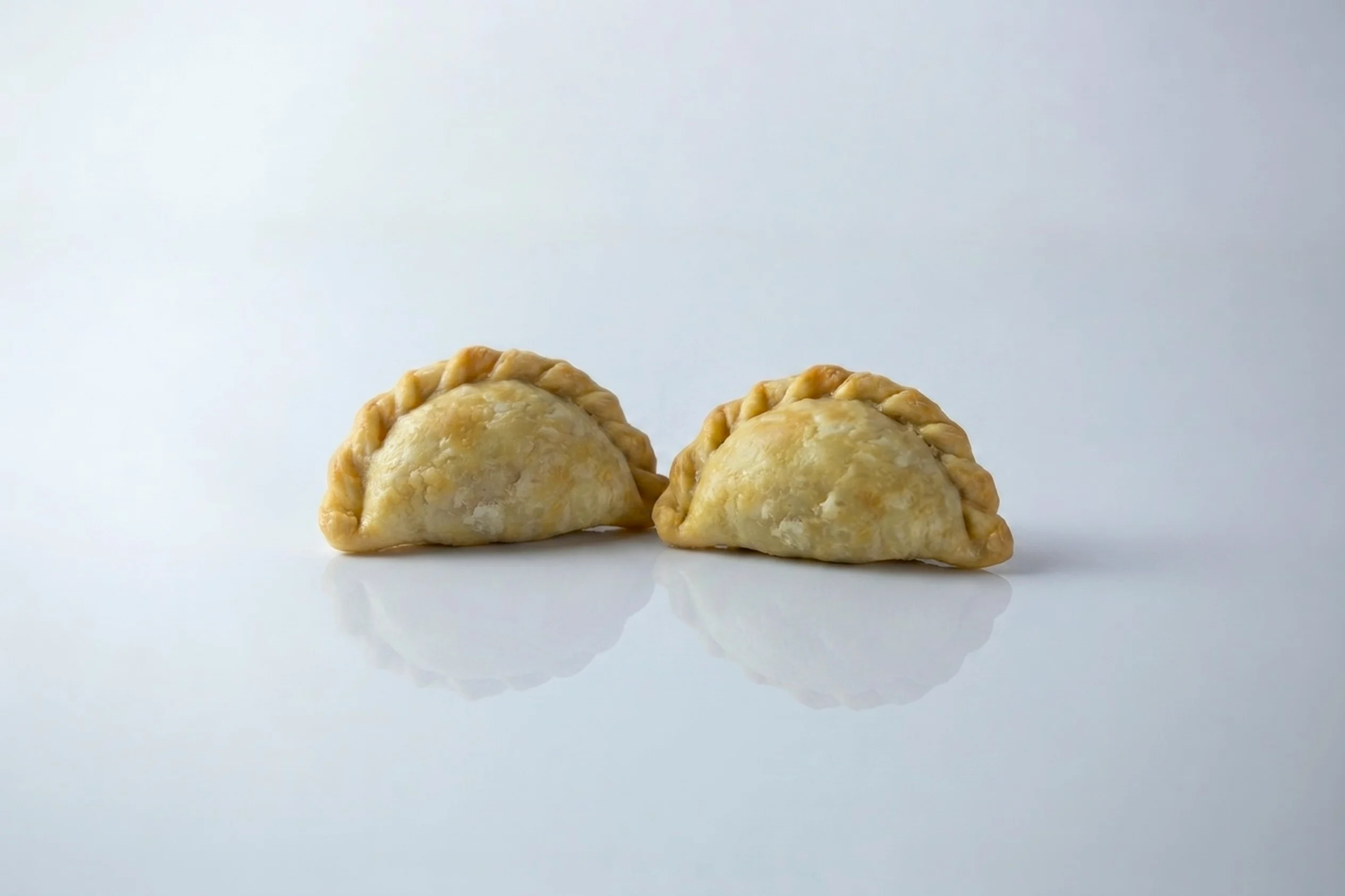 Two golden-baked empanadas with crimped edges on a white reflective surface against a light grey background.