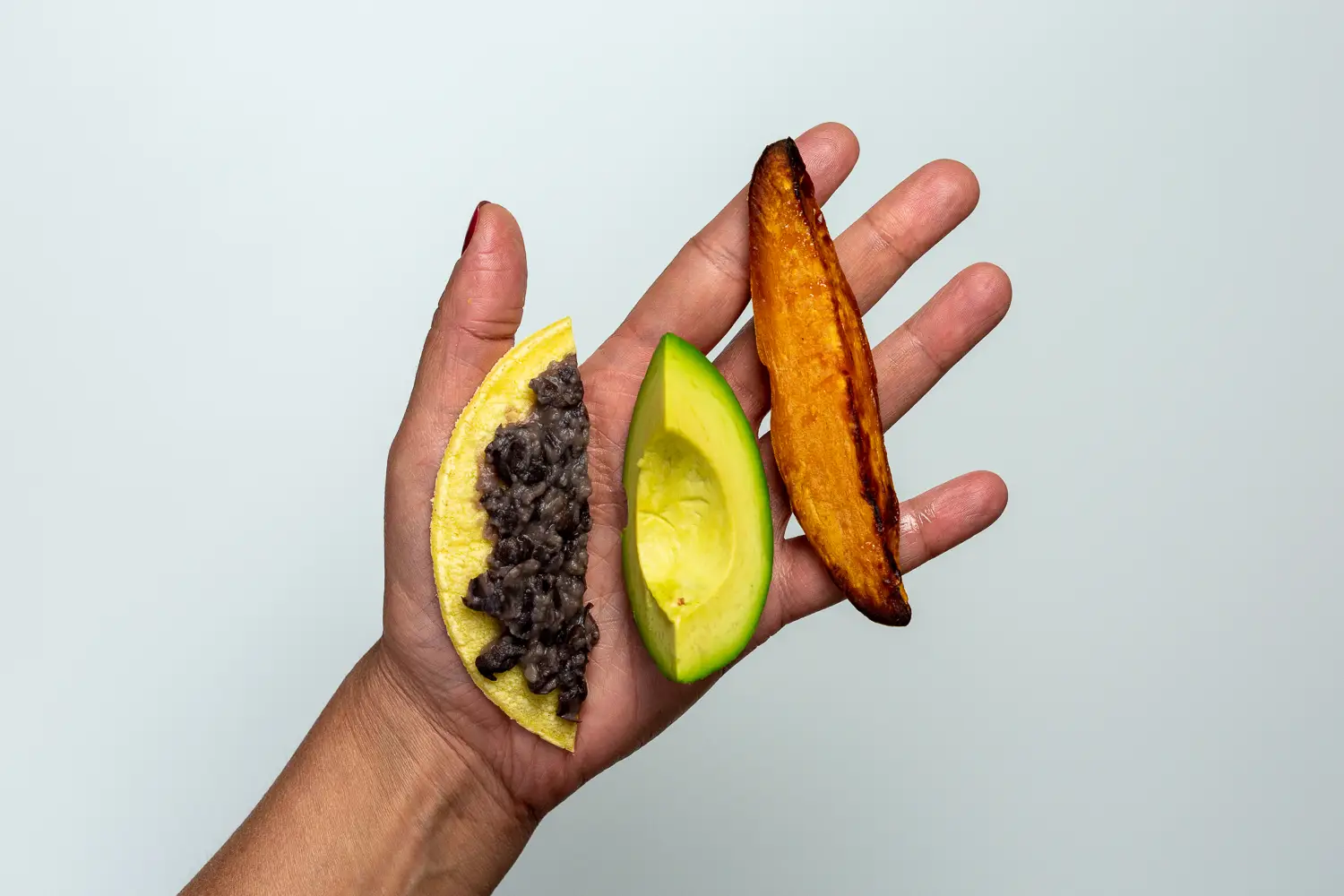 a photograph of a hand holding a strip of corn tortilla spread with mashed black beans, a wedge of avocado, and a spear of cooked sweet potato