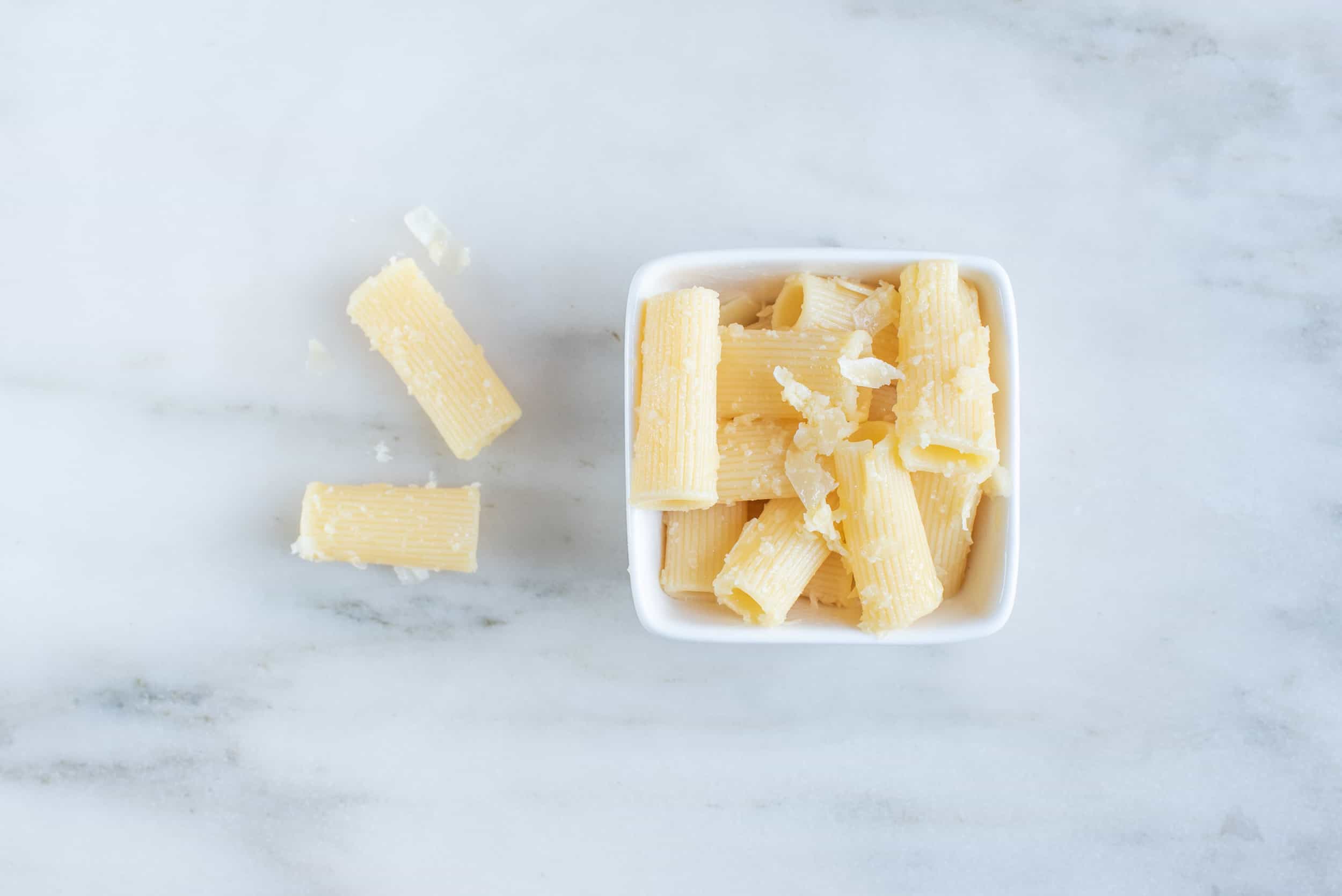 a square bowl filled with rigatoni pasta with two pieces of pasta next to the bowl on a white background