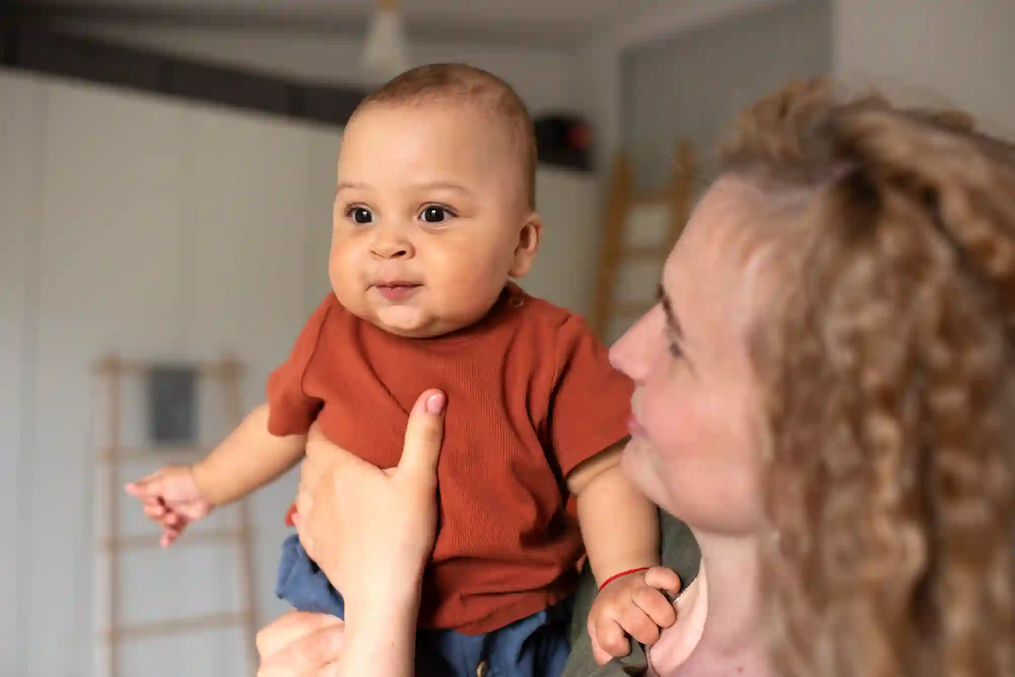 A woman with curly blonde hair holds up a baby in a rust-colored onesie, who looks alertly at the camera with a slight smile, in a bright home interior.