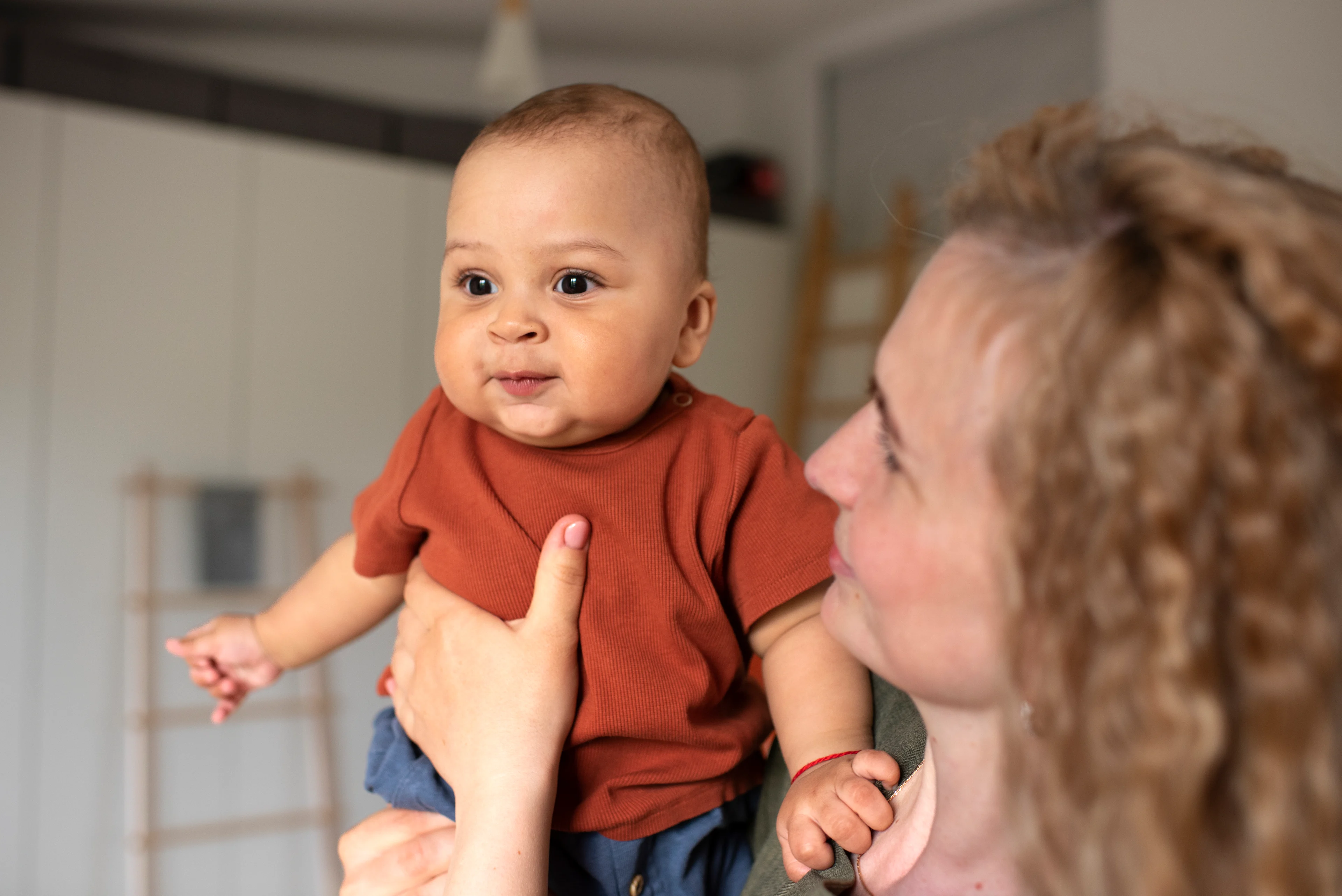 A woman with curly blonde hair holds up a baby in a rust-colored onesie, who looks alertly at the camera with a slight smile, in a bright home interior.