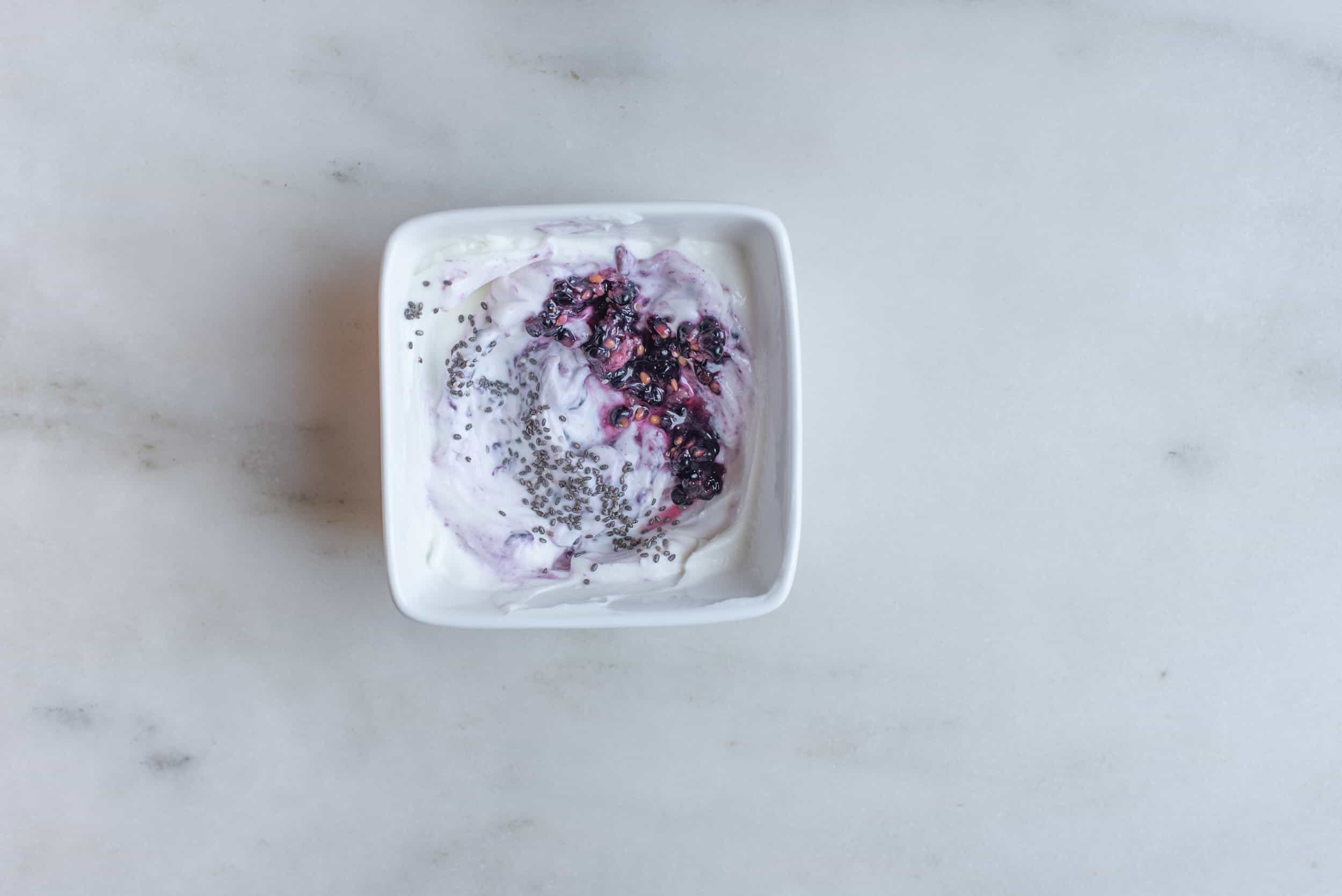 a white square bowl filled with yogurt, chia seed, mashed banana and mashed blackberry