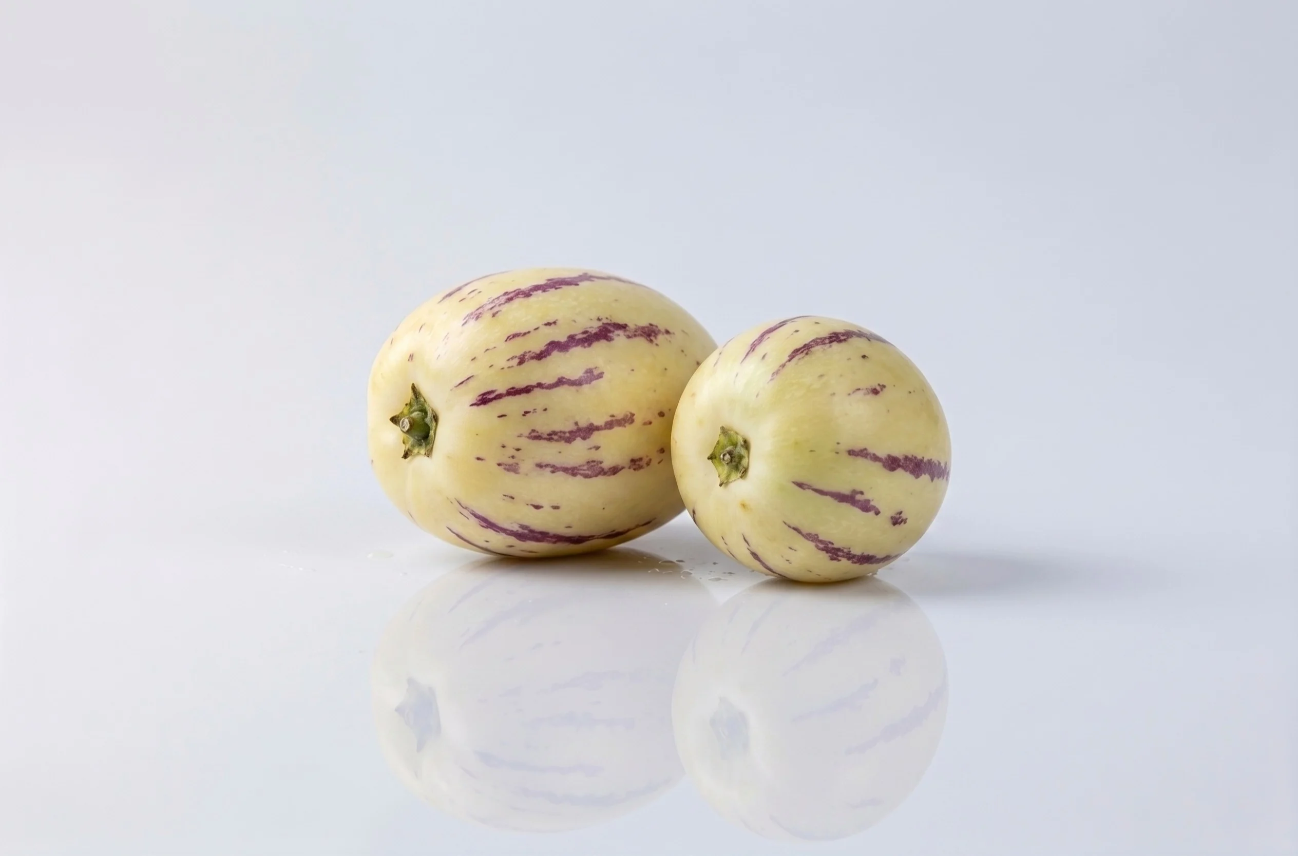 Two pepino melons with cream-colored skin and distinctive purple stripes on a white reflective surface against a light grey background, one larger than the other.