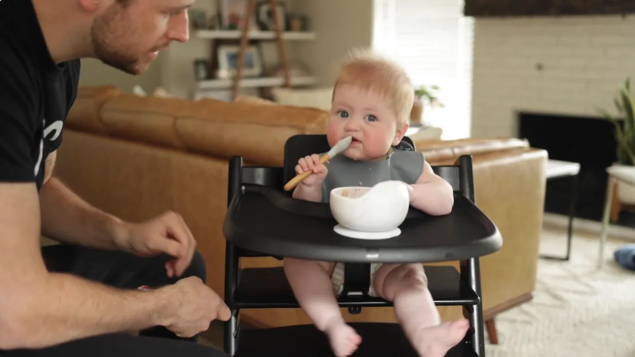 a photograph of a baby in a high chair with a bowl of food in front of them with his father looking on next to him