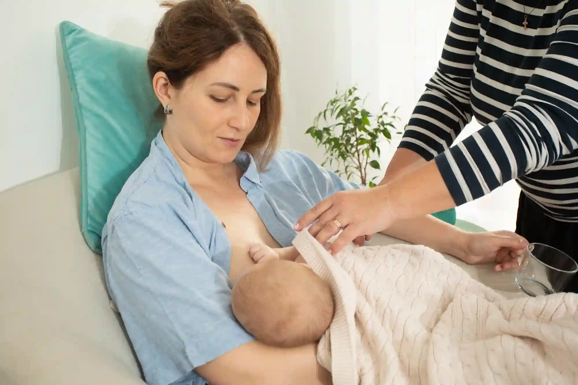 A mother breastfeeding her baby while reclining on a couch, with a healthcare provider or support person assisting her and offering a glass of water.