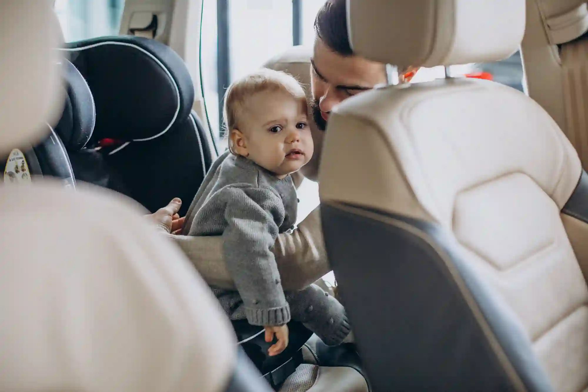 A parent carefully secures a baby in a car seat inside a vehicle. The baby, wearing a gray sweater, looks curious and slightly amused.