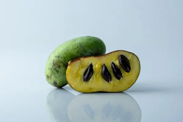 a whole pawpaw behind a pawpaw cut in half to show the large, black seeds inside