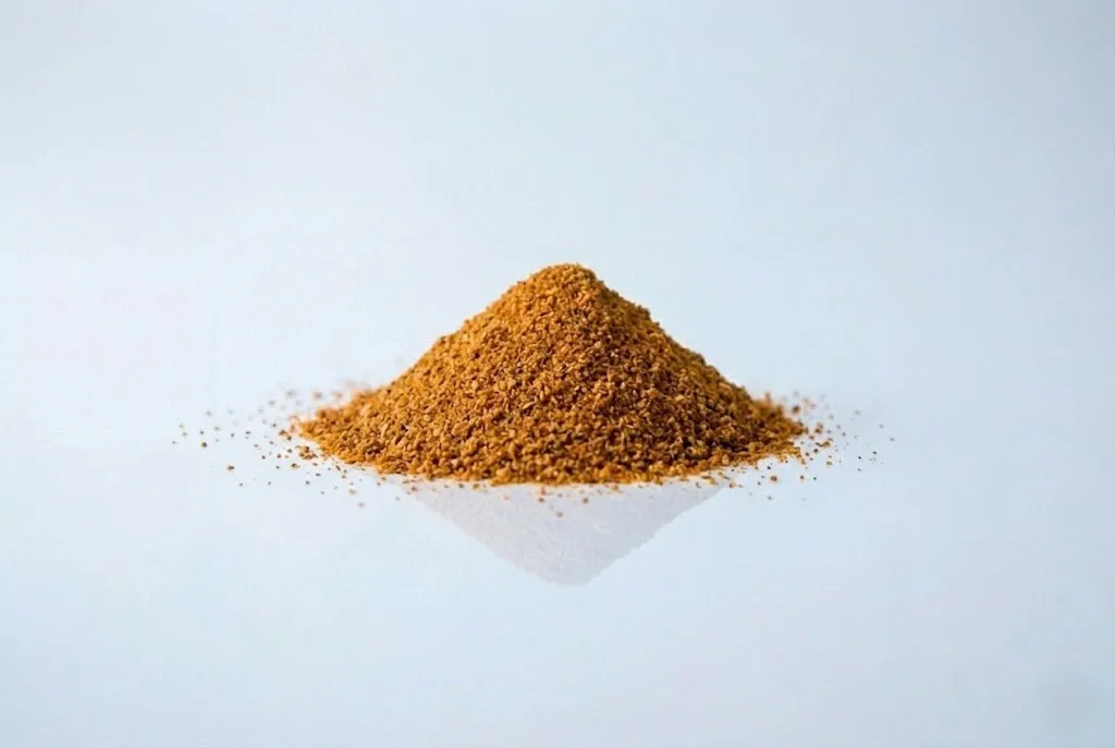 A small mound of golden-orange adobo seasoning powder on a white reflective surface against a light grey background.