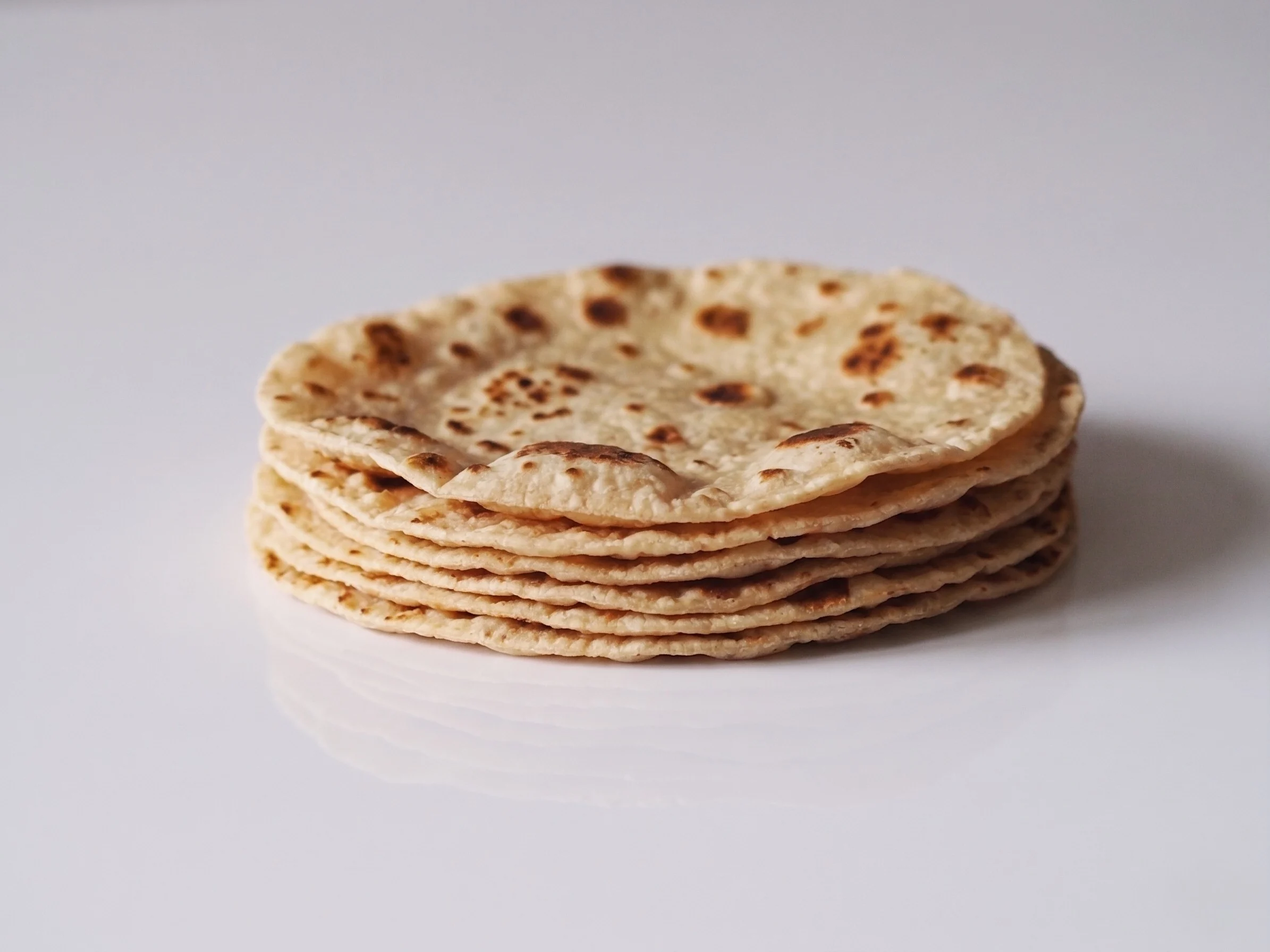 A stack of freshly cooked roti with golden-brown char marks on a white reflective surface against a light grey background.