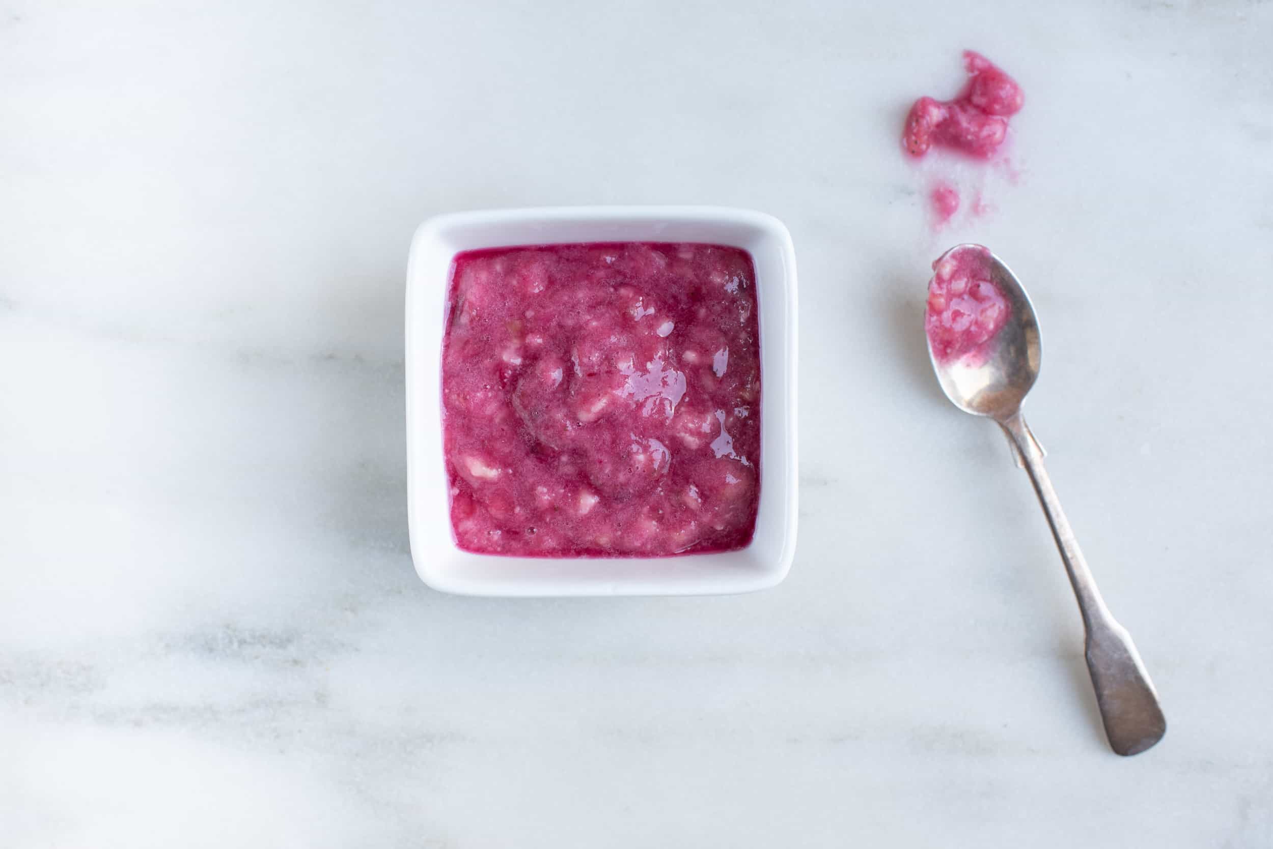 a square white bowl filled with pink prickly pear apple sauce next to a metal spoon that holds a little of the sauce