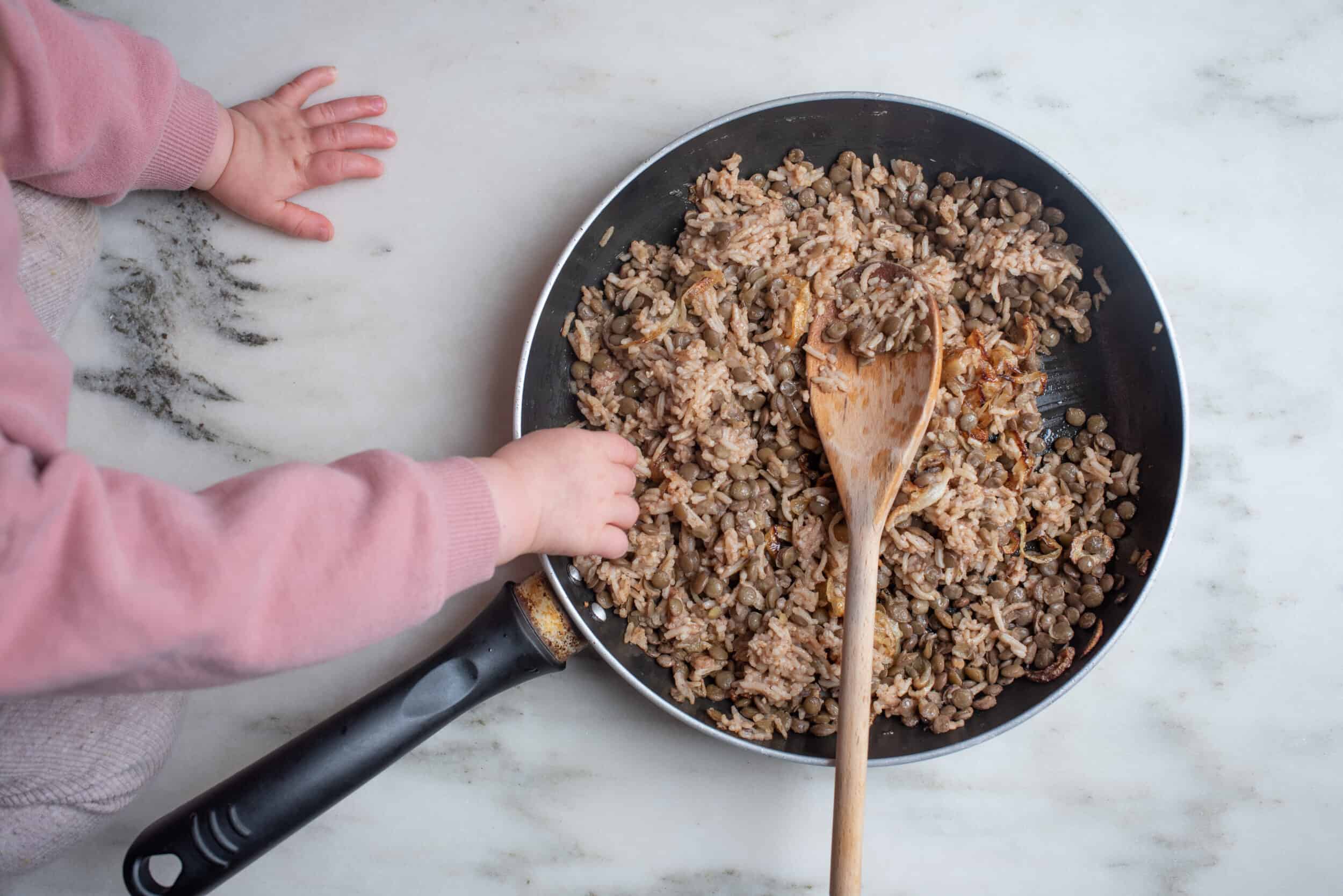 baby's hand reaching into a frying pan filled with lentils, sitting on a countertop with a wooden spoon in the pan