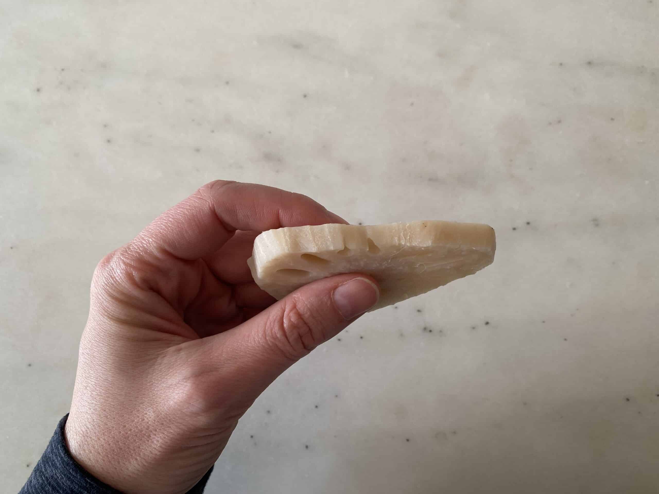a hand holding a thin cooked slice of lotus root to show the slice's thickness