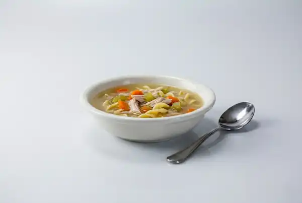a photograph of a shallow white bowl with chicken noodle soup in it and a spoon lying next to the bowl