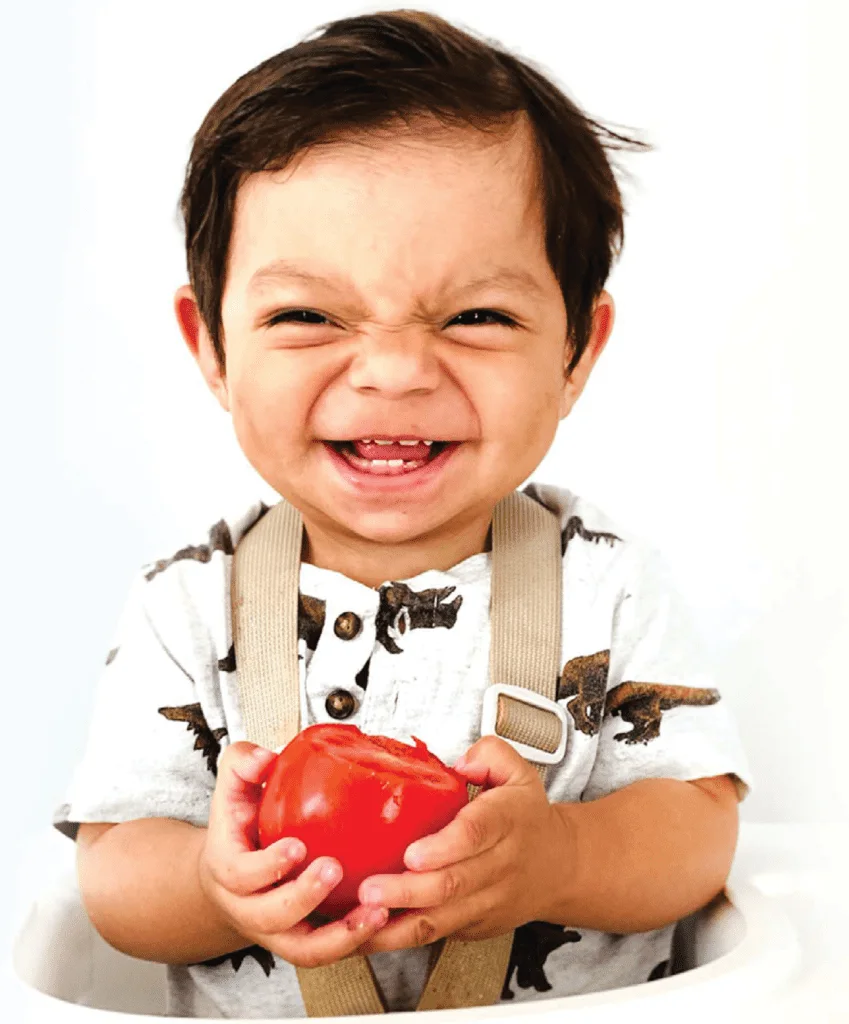 toddler strapped to a high chair smiling and holding a tomato with both hands