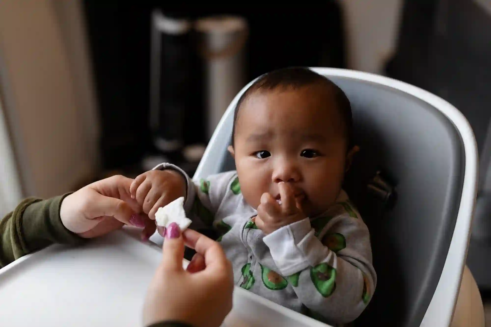 A baby in an avocado-print onesie sitting in a white high chair, eating while an adult offers a piece of soft white food, with the baby's hand also reaching toward their mouth.