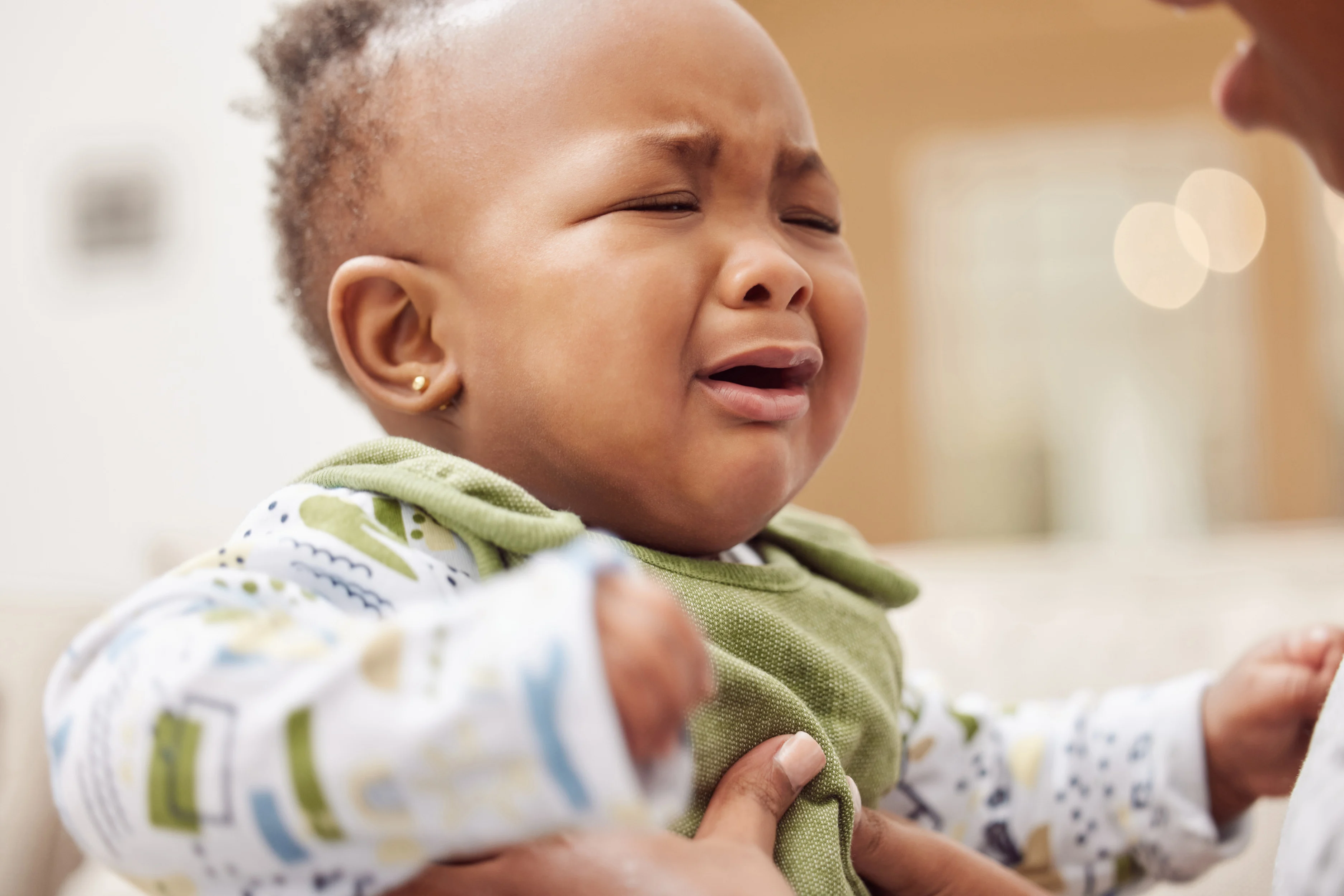 A crying baby wearing a green bib and patterned shirt is being comforted by a person. The scene conveys a sense of distress and care.