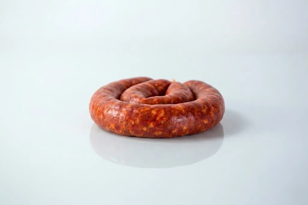 A coiled raw chorizo sausage link with a deep red color on a white reflective surface against a light grey background.