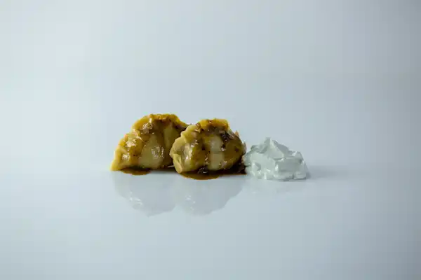 Two brown dumplings glazed with sauce, paired with a dollop of white cream on a smooth, white surface. A minimalist and appetizing setup.
