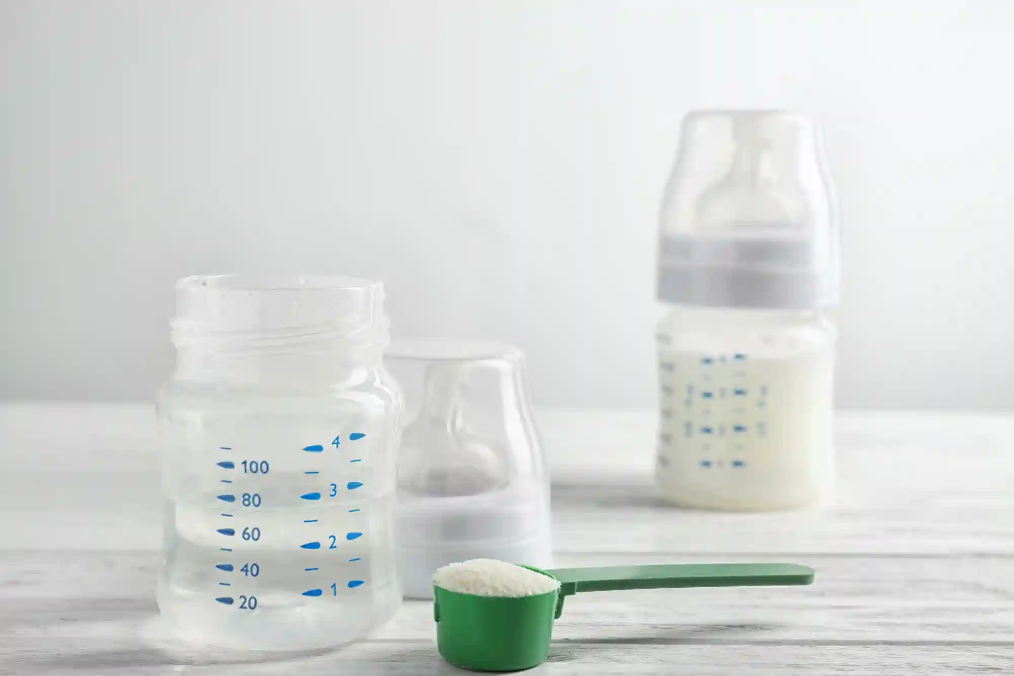 A green measuring scoop filled with powdered infant formula in front of an empty baby bottle with measurement markings, with a second bottle filled with prepared formula in the background.