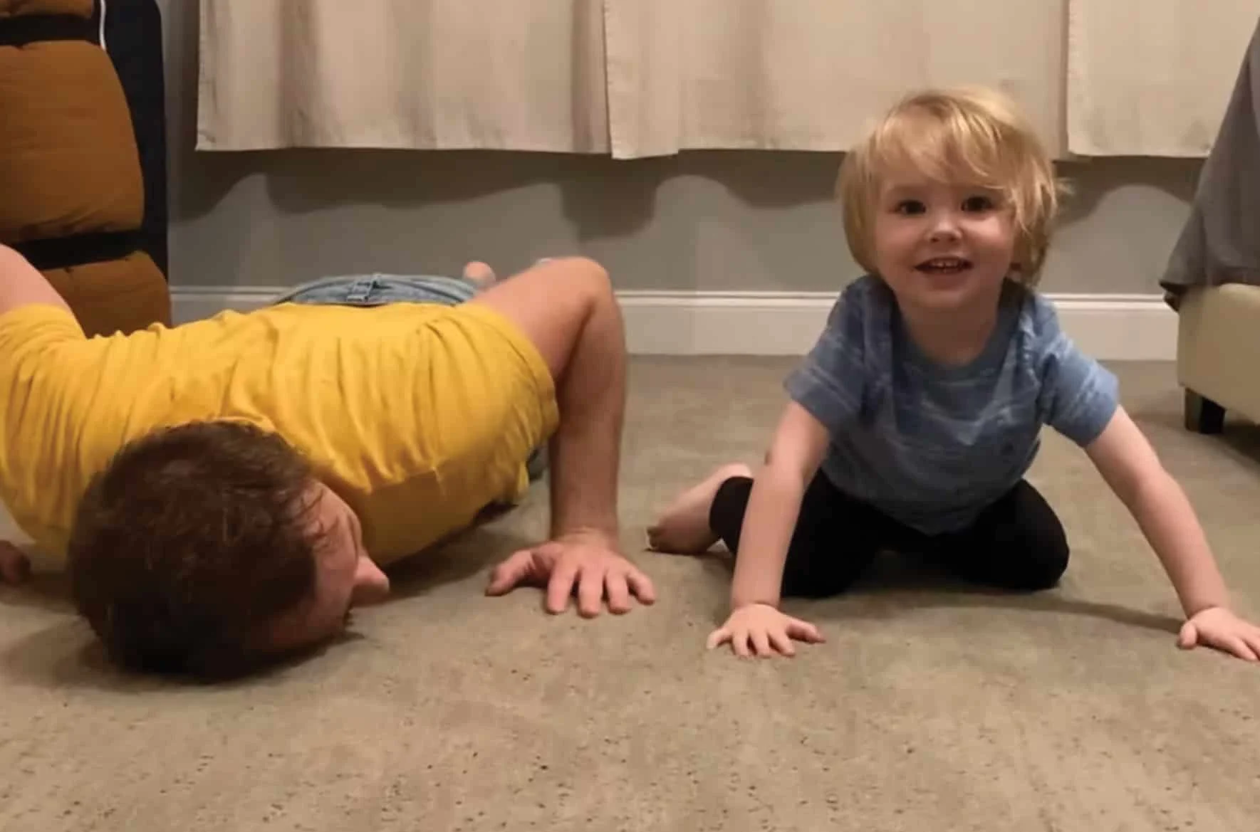 un niño pequeño sonriendo sentado en el piso a lado de un hombre acostado boca abajo en el piso