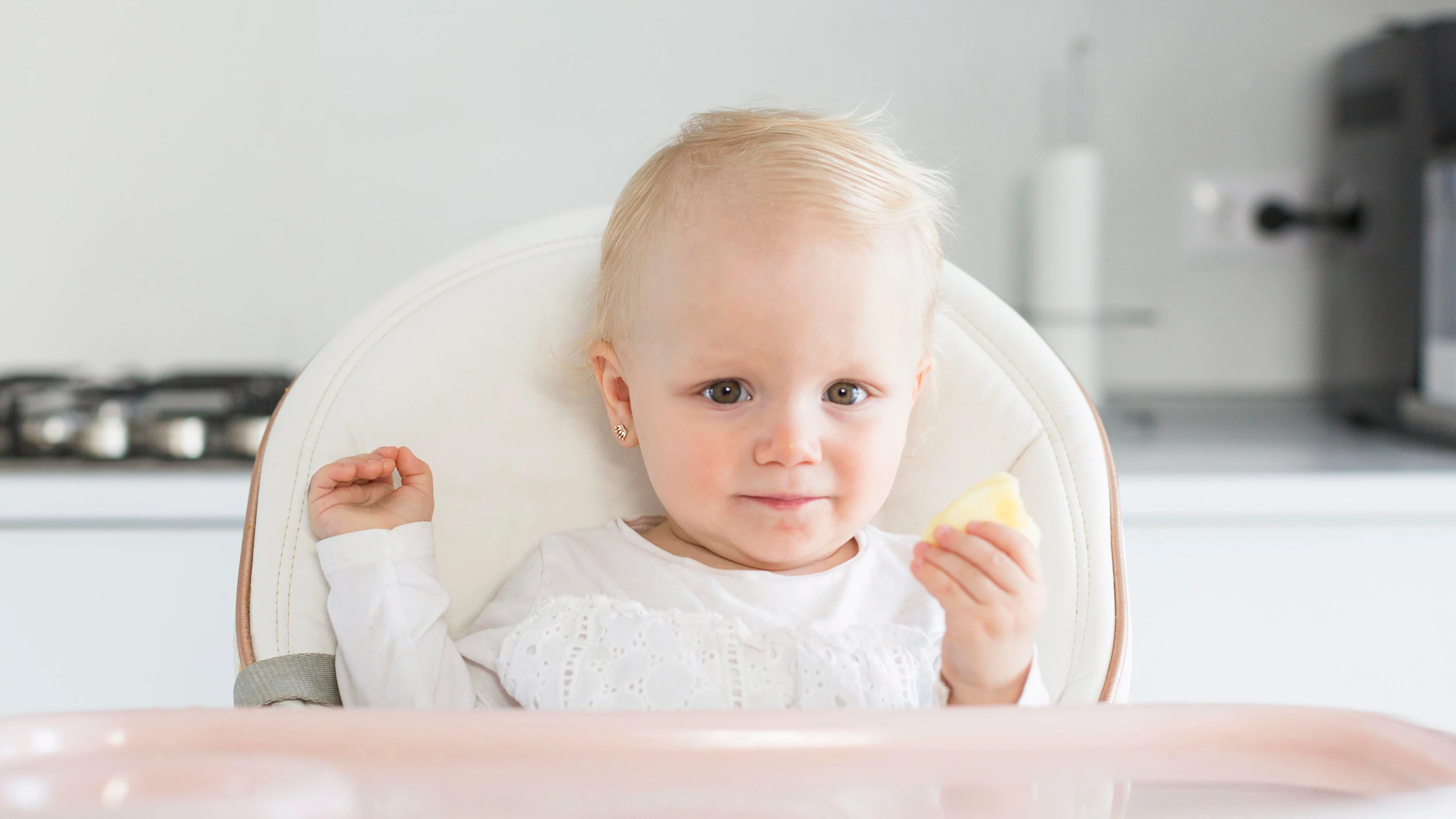 A blonde baby girl sitting in a white high chair, looking at the camera while holding a small piece of food, with a modern kitchen in the blurred background.