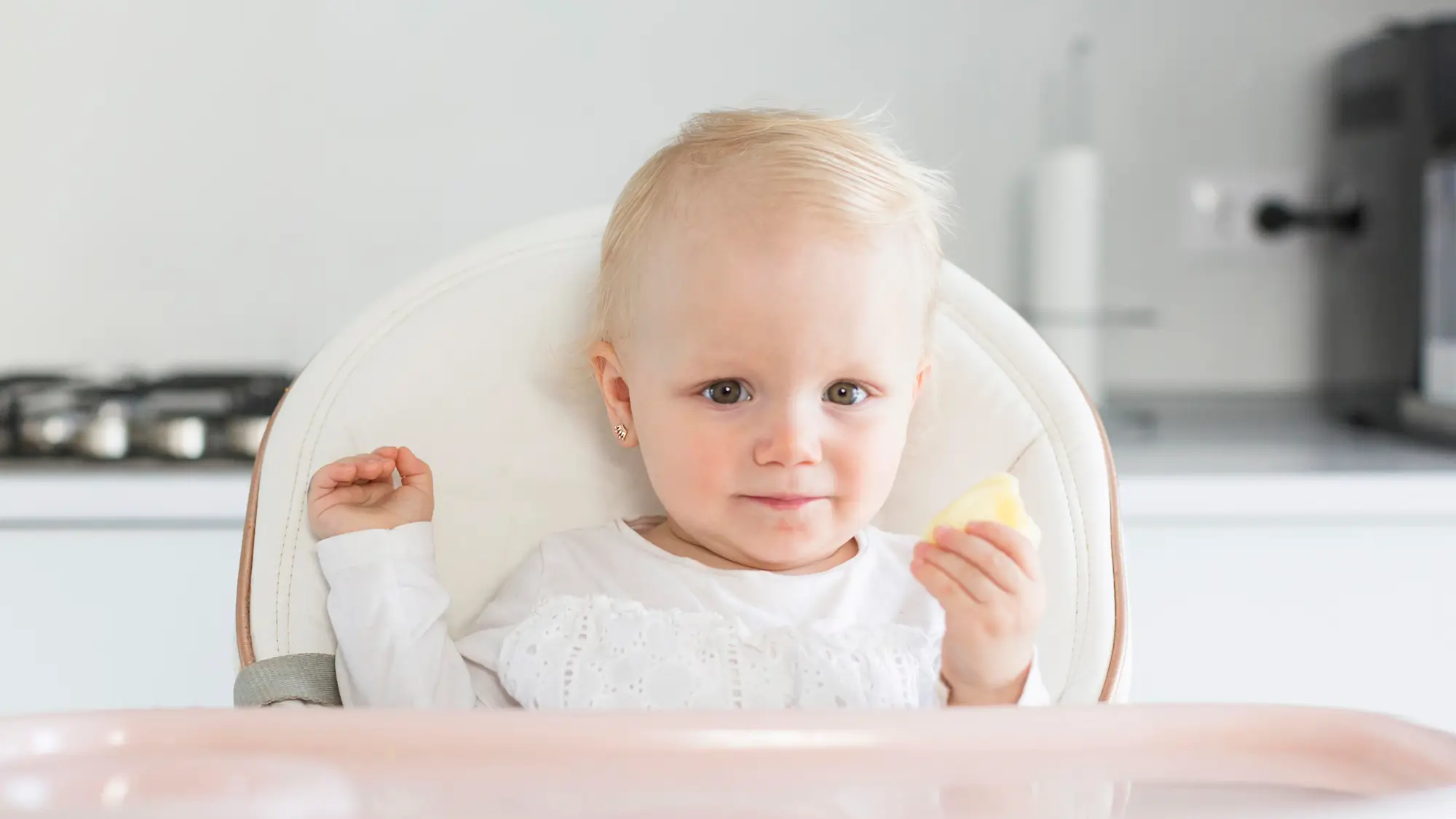 solids-and-reflux A blonde baby girl sitting in a white high chair, looking at the camera while holding a small piece of food, with a modern kitchen in the blurred background.