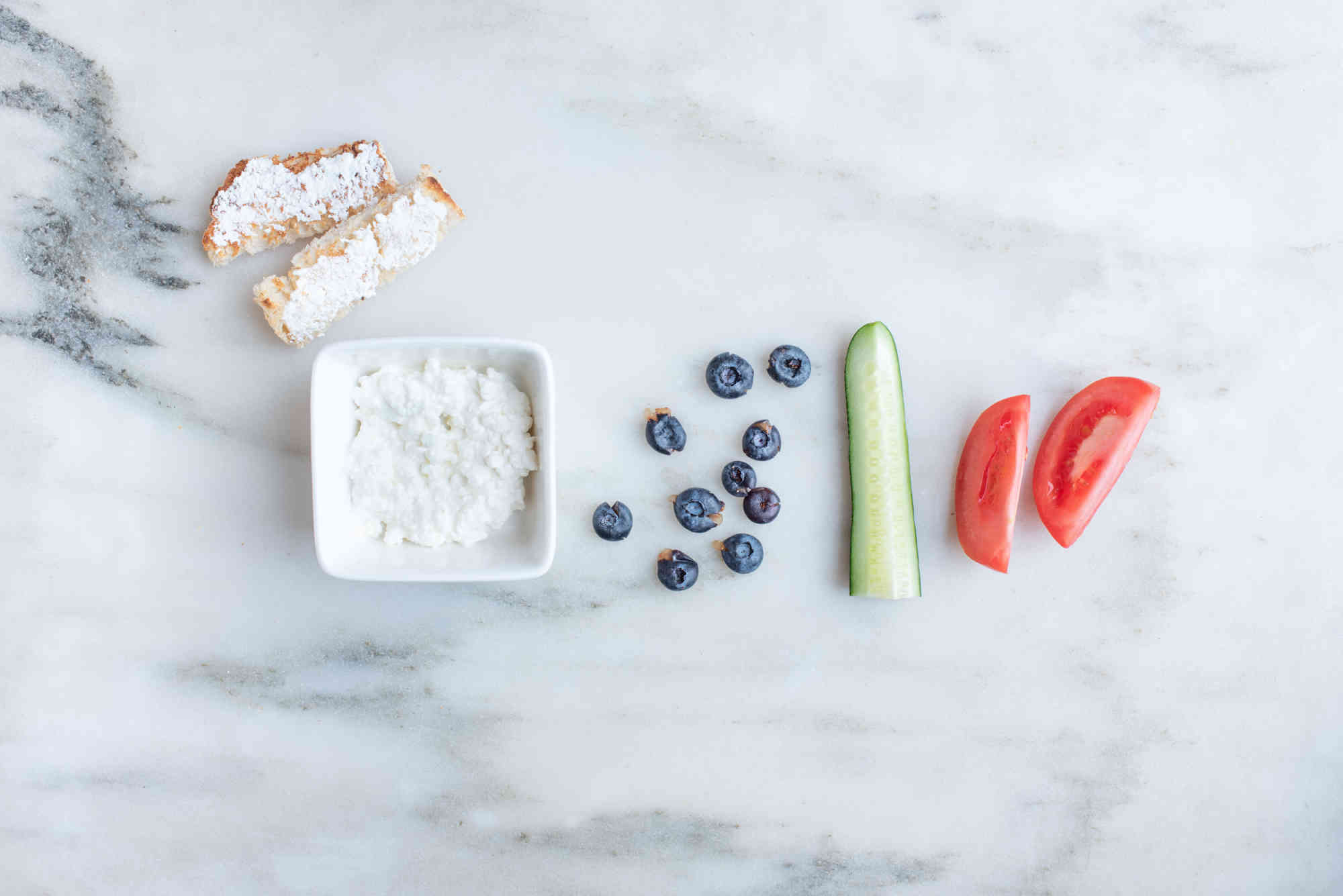 a square white bowl filled with cottage cheese in between two toast strips spread with cottage cheese, and 10 flattened blueberries, a cucumber spear, and two large tomato wedges