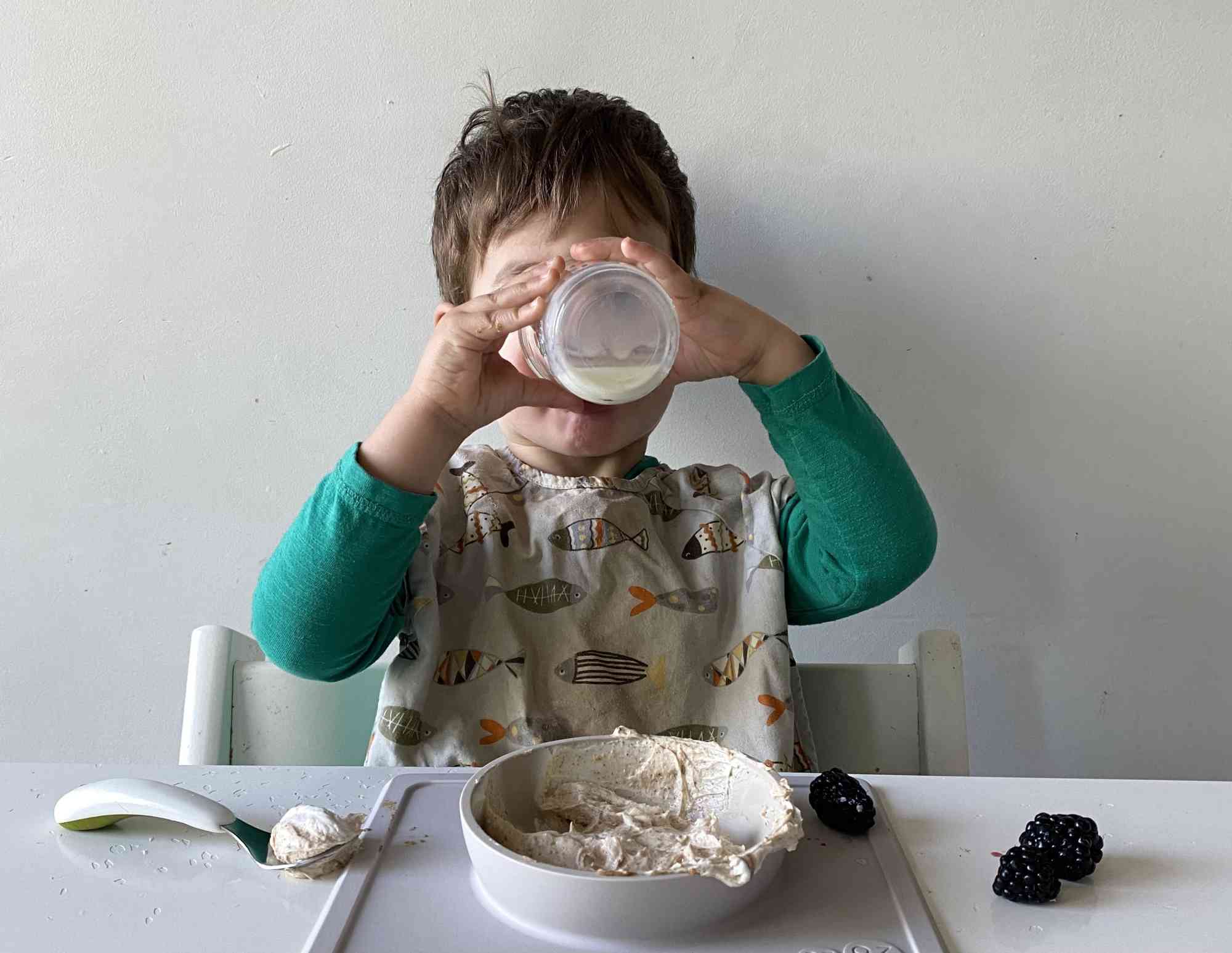 Toddler sitting in chair at a table, with a glass of milk up to his mouth. 