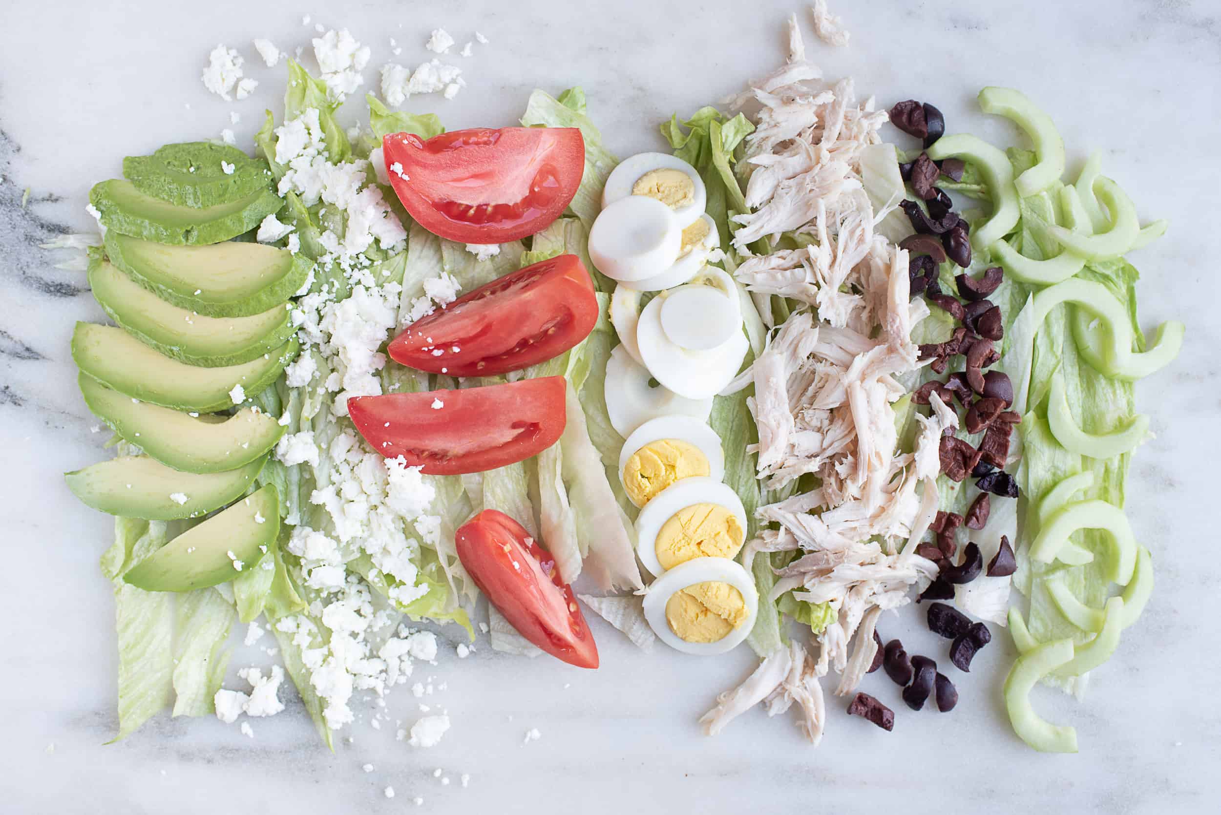 iceberg lettuce leaves sliced into ribbons, sliced hardboiled eggs, tomato wedges, shredded chicken, sliced avocado, half moon slices of cucumber, sliced olives, and crumbled goat cheese, all arranged in columns by food type, on a countertop