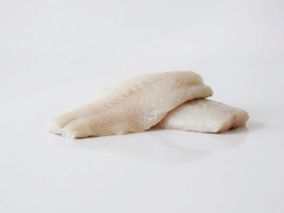 Two raw white fish fillets stacked on a white reflective surface against a light grey background.
