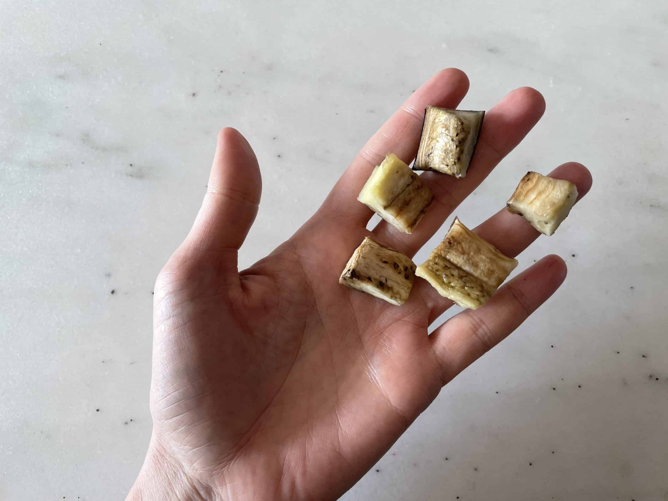 a hand holding five bite-sized pieces of cooked eggplant for babies 9 months+