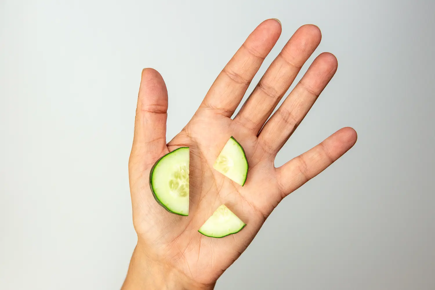a photograph of a hand holding three thin slices of cucumber, one cut in half into a half-moon and one cut in quarters