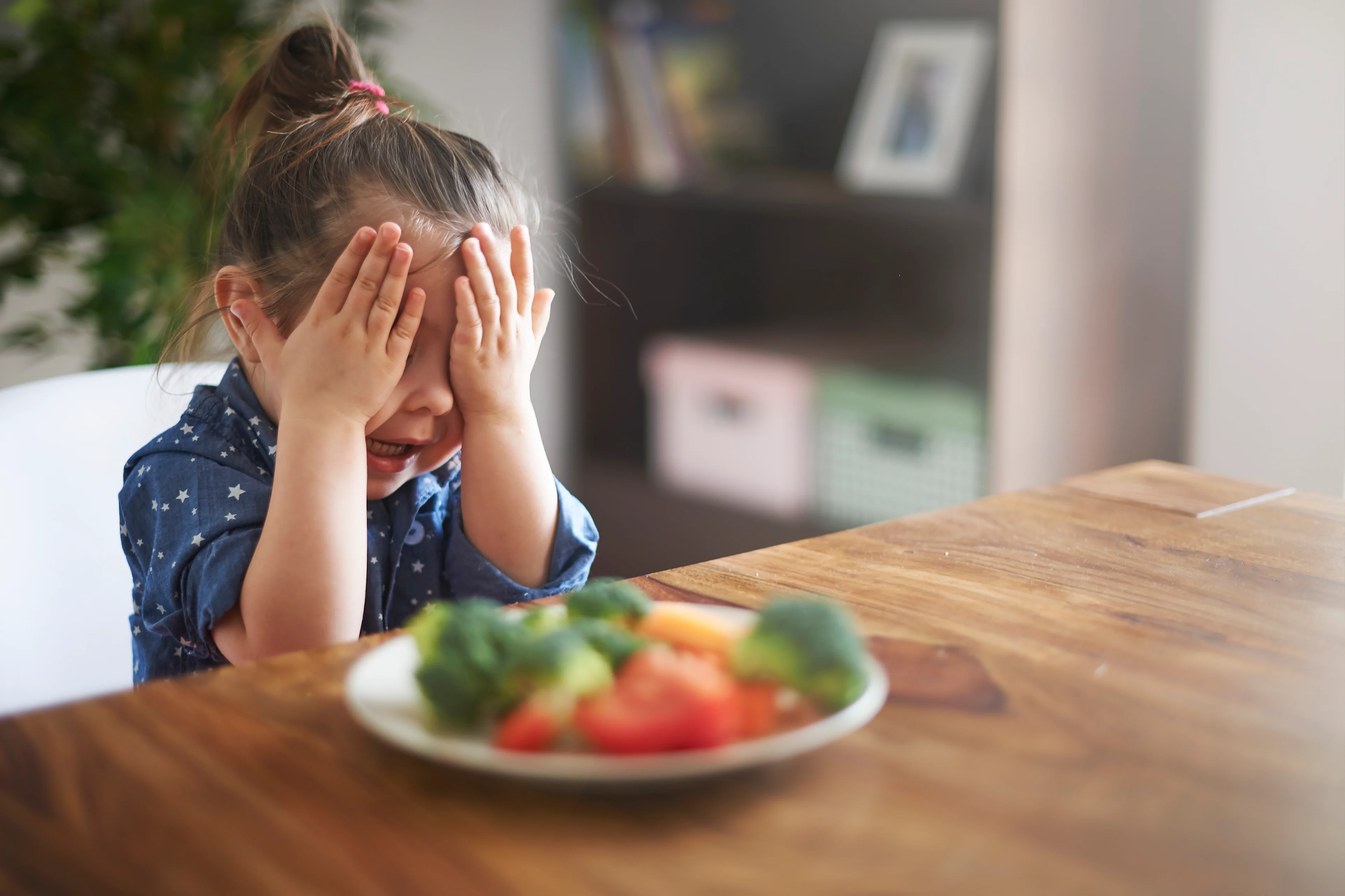 A little girl in a blue star-print top covers her face with both hands in protest in front of a plate of broccoli, tomatoes, and other vegetables on a wooden table.