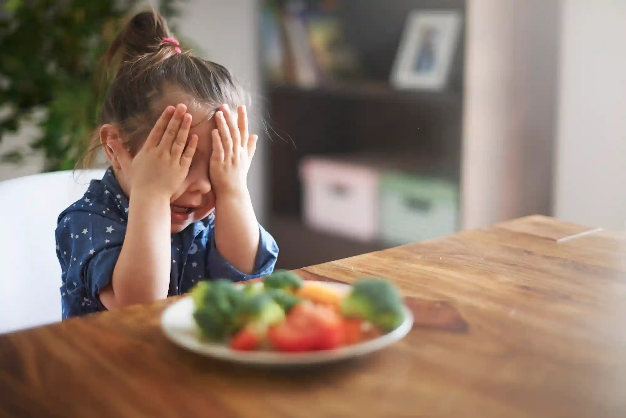 Picky Eater - Little Girl A little girl in a blue star-print top covers her face with both hands in protest in front of a plate of broccoli, tomatoes, and other vegetables on a wooden table.