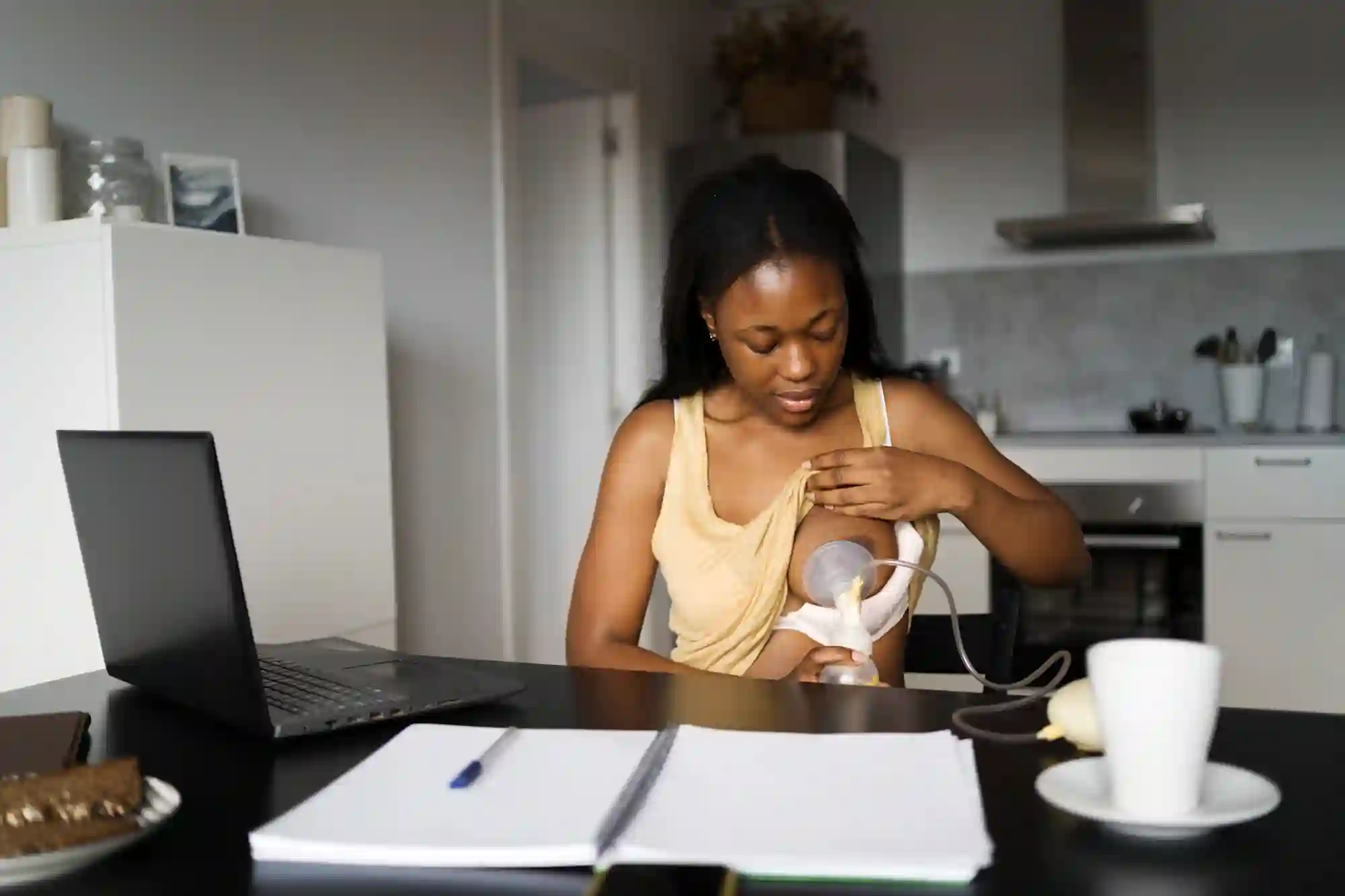 A woman in a kitchen uses a breast pump while seated at a table with a laptop, notebook, and coffee. The scene conveys multitasking and focus.