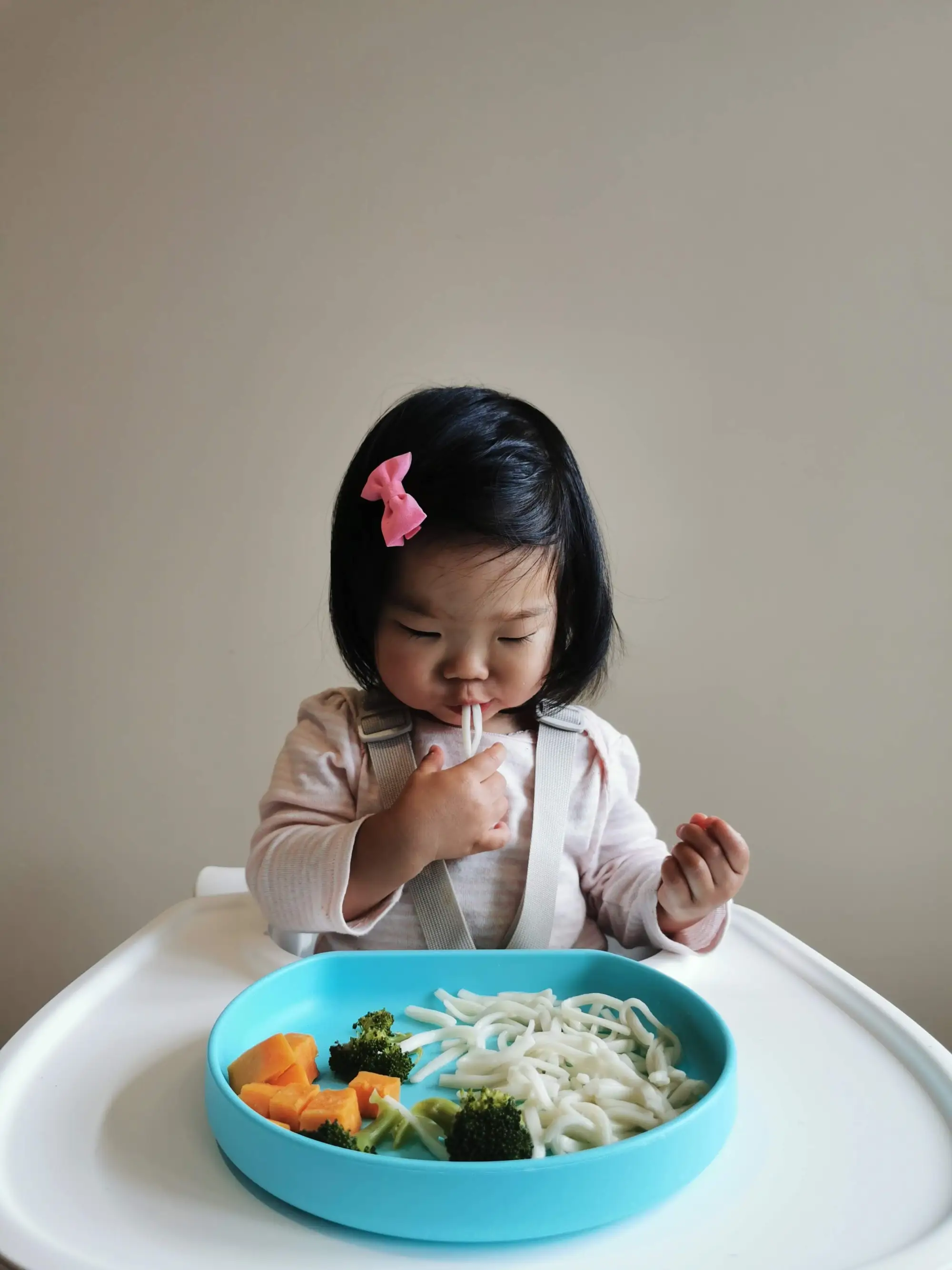 a toddler in a high chair eats noodles and vegetables from a blue bowl